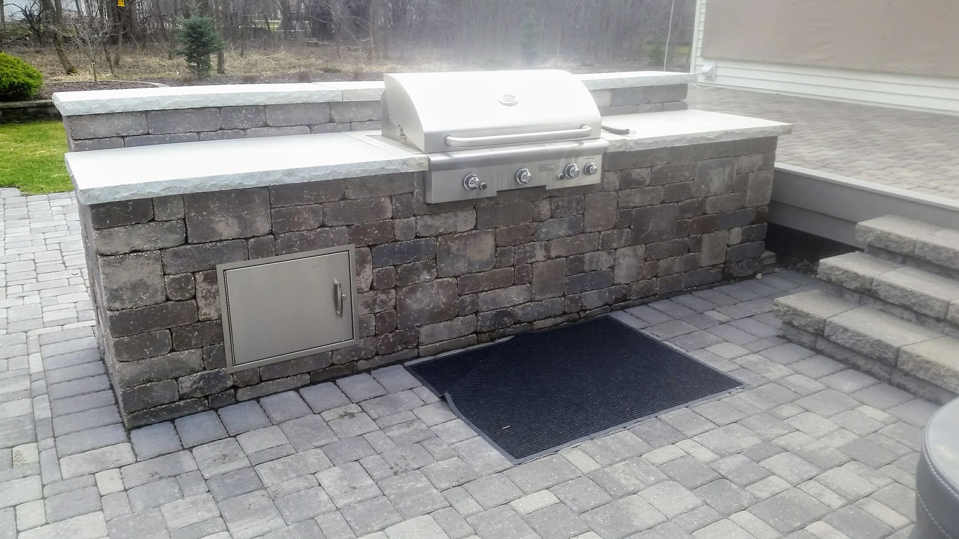 An outdoor stone kitchen island with a stainless steel grill and cabinet, situated on a gray paver patio.