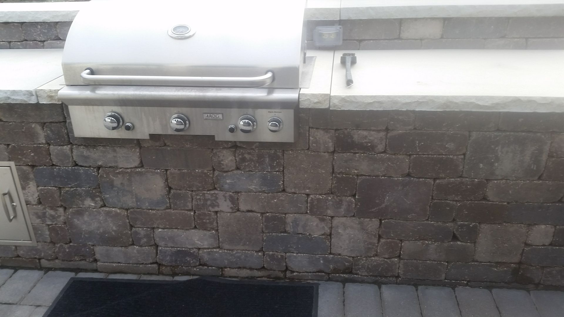Stainless steel outdoor grill built into a rustic stone kitchen island on a paved patio.