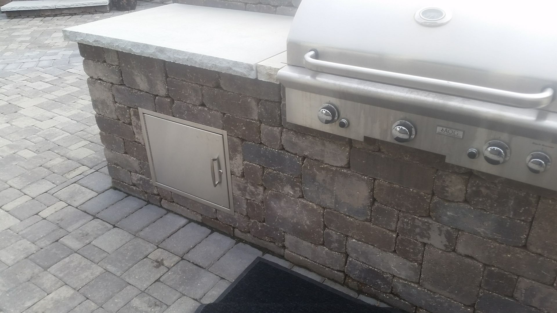 An outdoor stone kitchen island with a stainless steel grill and access door on a paved patio.