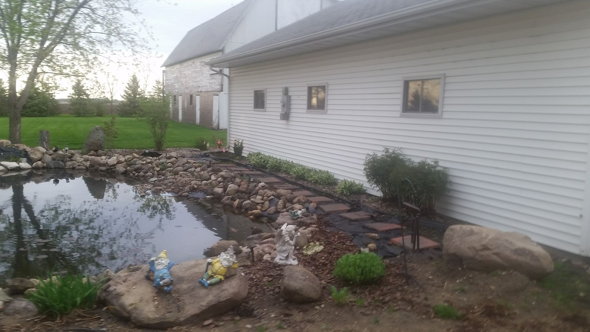 A stone-lined pond sits beside a white-sided building with garden beds, a path, and lawn ornaments in a grassy yard.