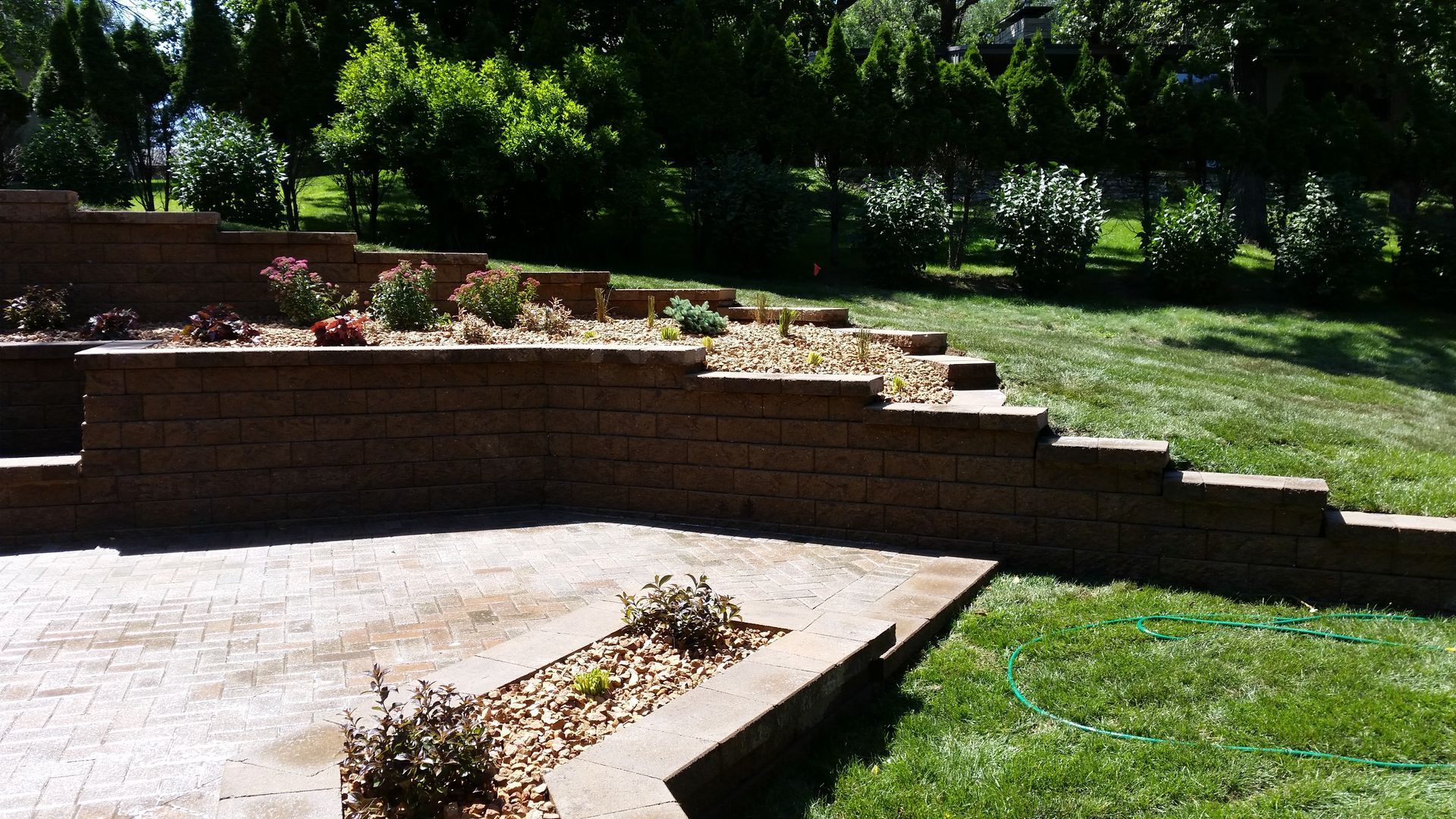 A multi-tiered brown stone retaining wall borders a stone patio and garden bed, set against a green lawn and trees.