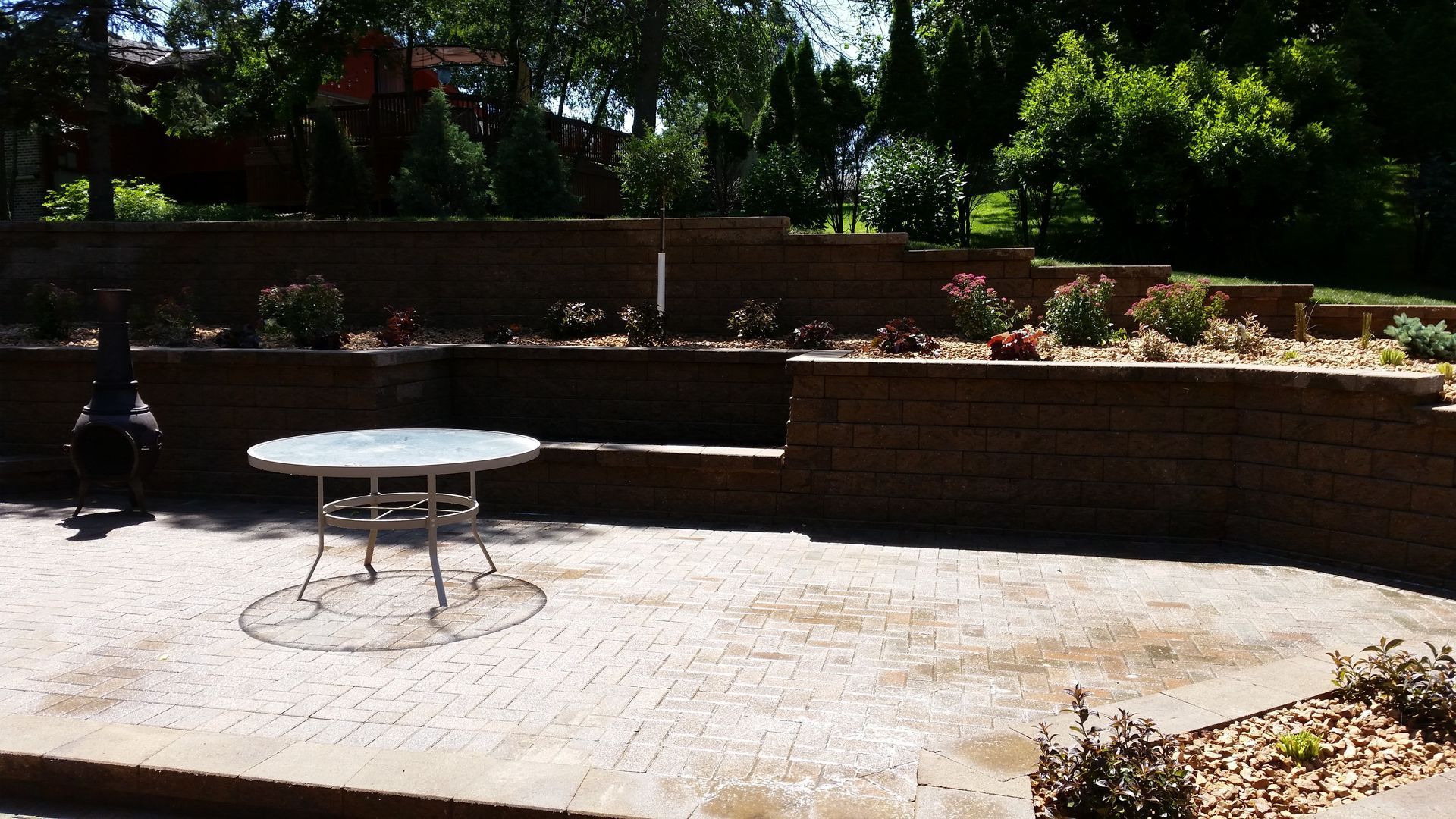 A patio with a round glass table, surrounded by multi-tiered stone retaining walls and landscaping in a backyard.