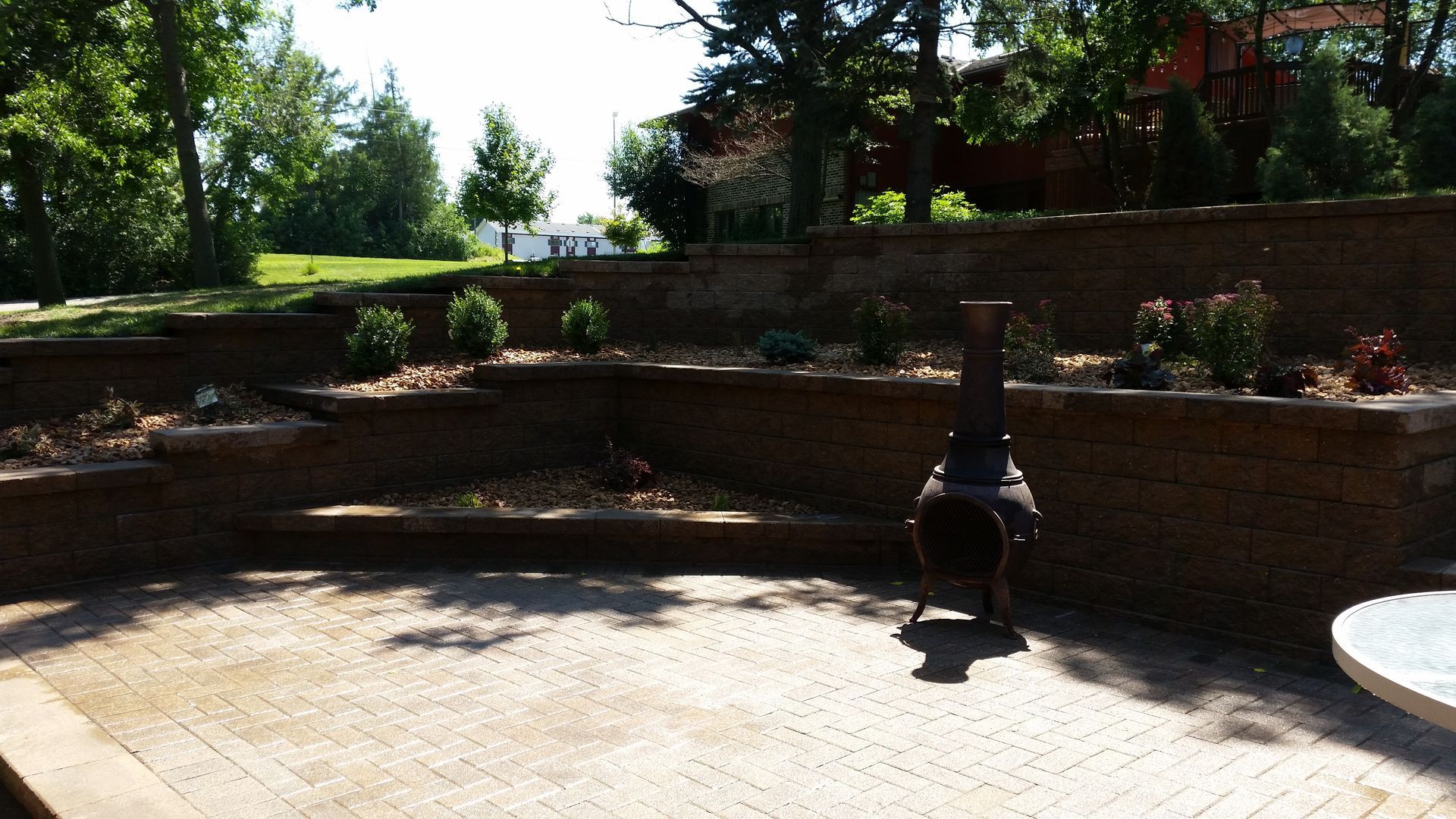 A stone patio with tiered brick retaining walls, gravel landscaping, small bushes, and a metal outdoor fireplace.