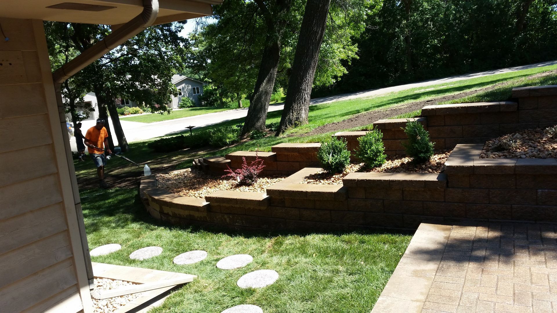 A person in an orange shirt works in a backyard with a tiered retaining wall, landscaping, and a paver stone pathway.