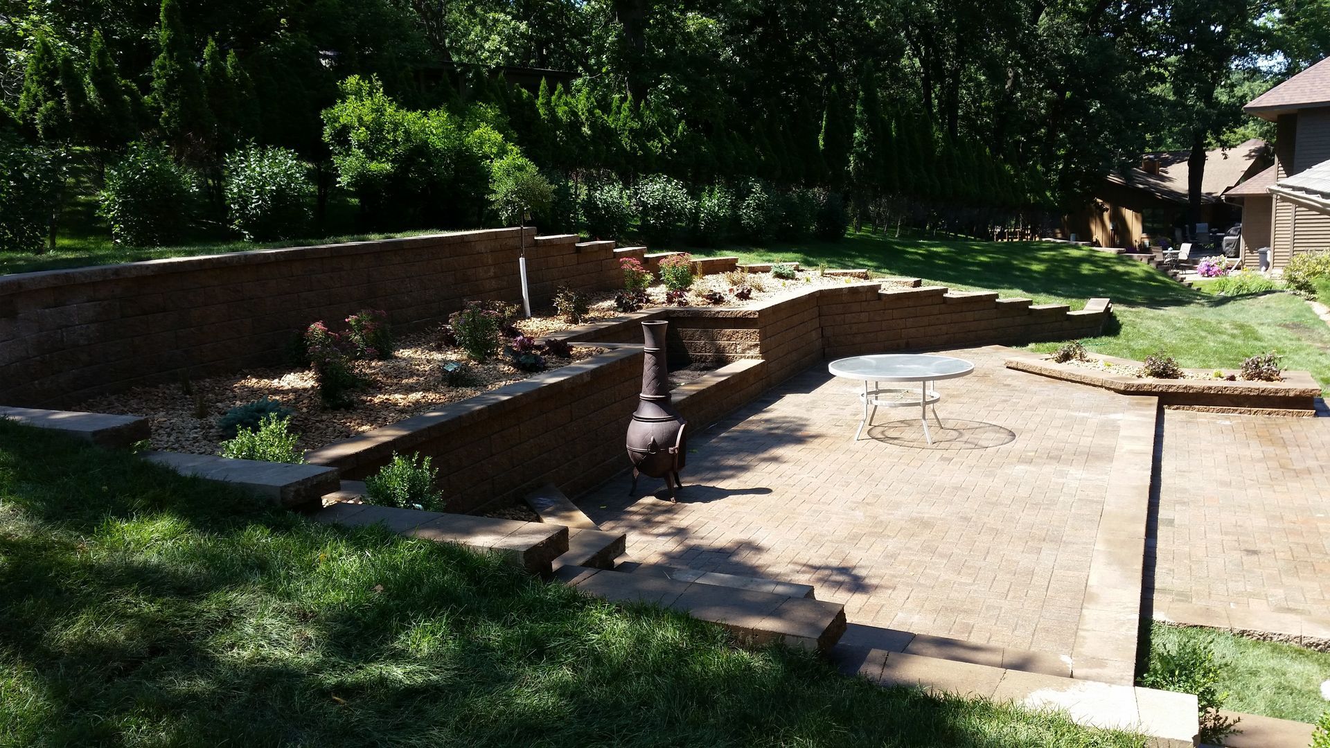 A terraced backyard landscape with brick retaining walls, a stone patio, a small fire pit, and scattered plants.