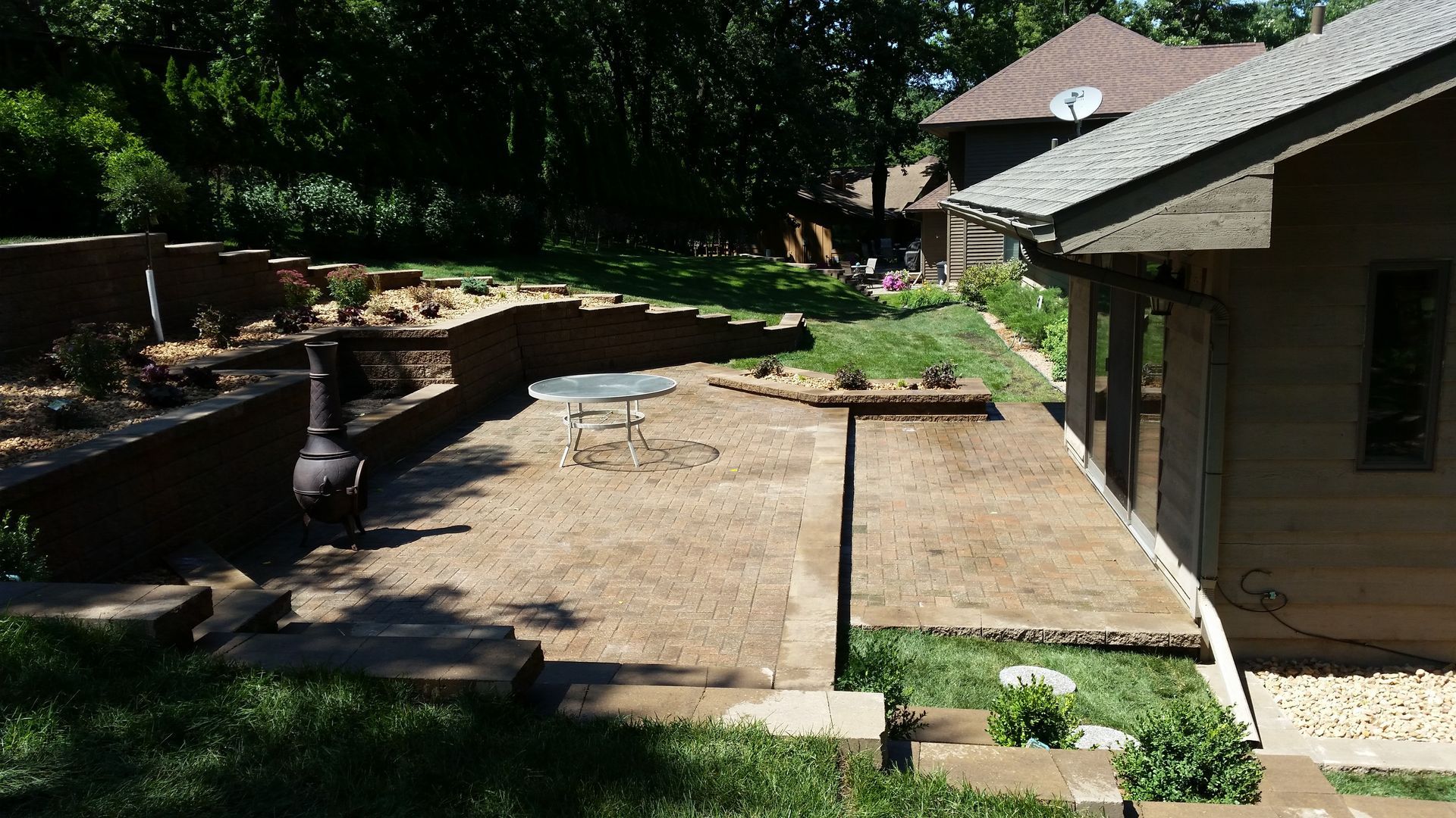 A backyard features a gravel patio with a table next to a house, surrounded by tiered retaining walls and green lawns.