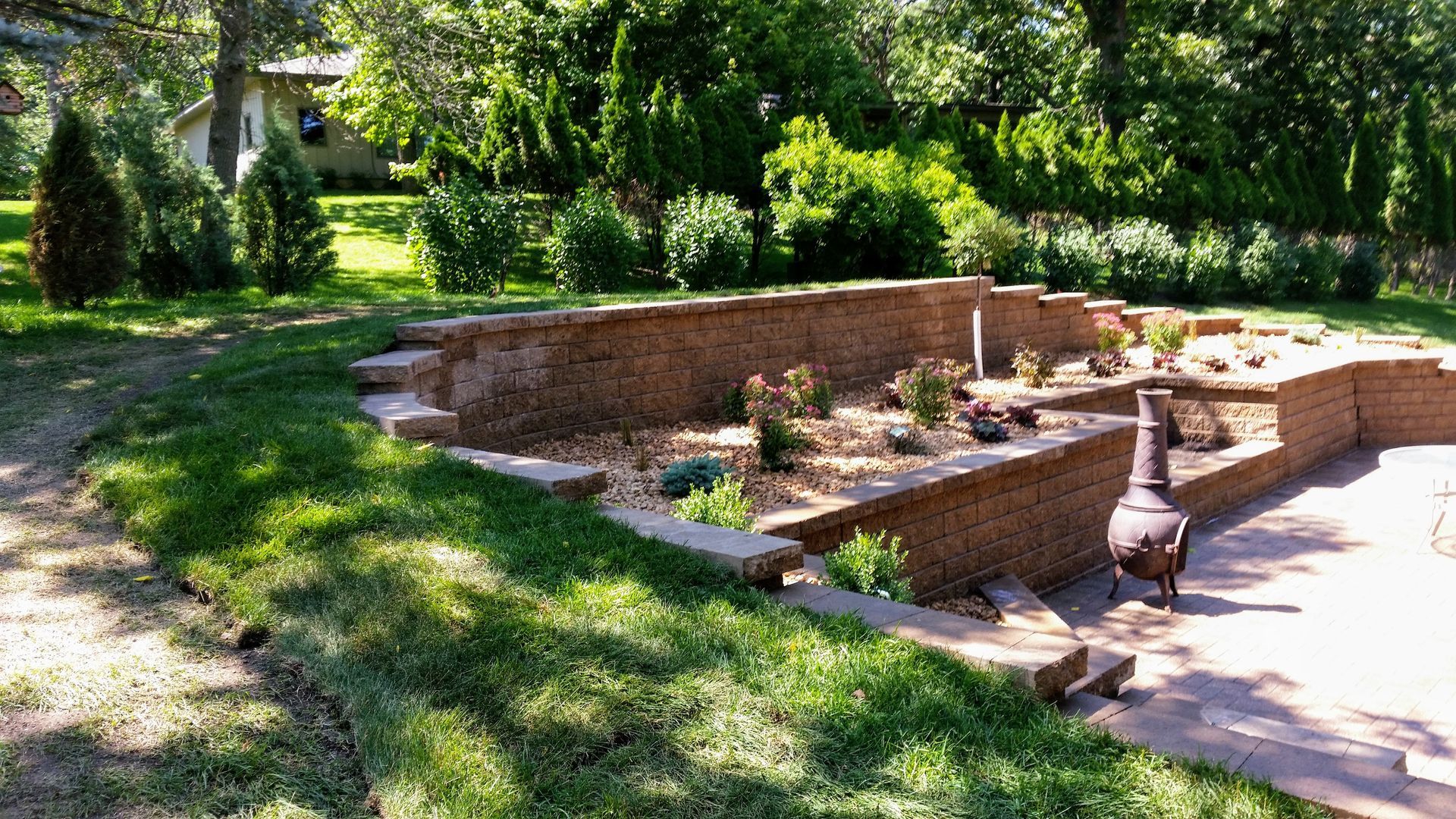A multi-tiered brick retaining wall features a landscaped bed with gravel and small plants next to a patio and fire pit.