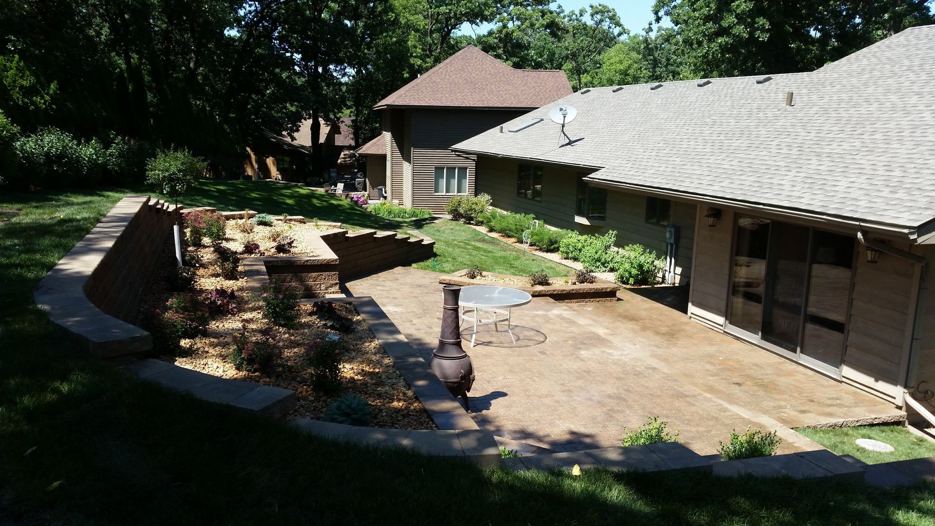 A backyard stone patio with a curved retaining wall, rock garden, and fire pit next to a house under a clear blue sky.
