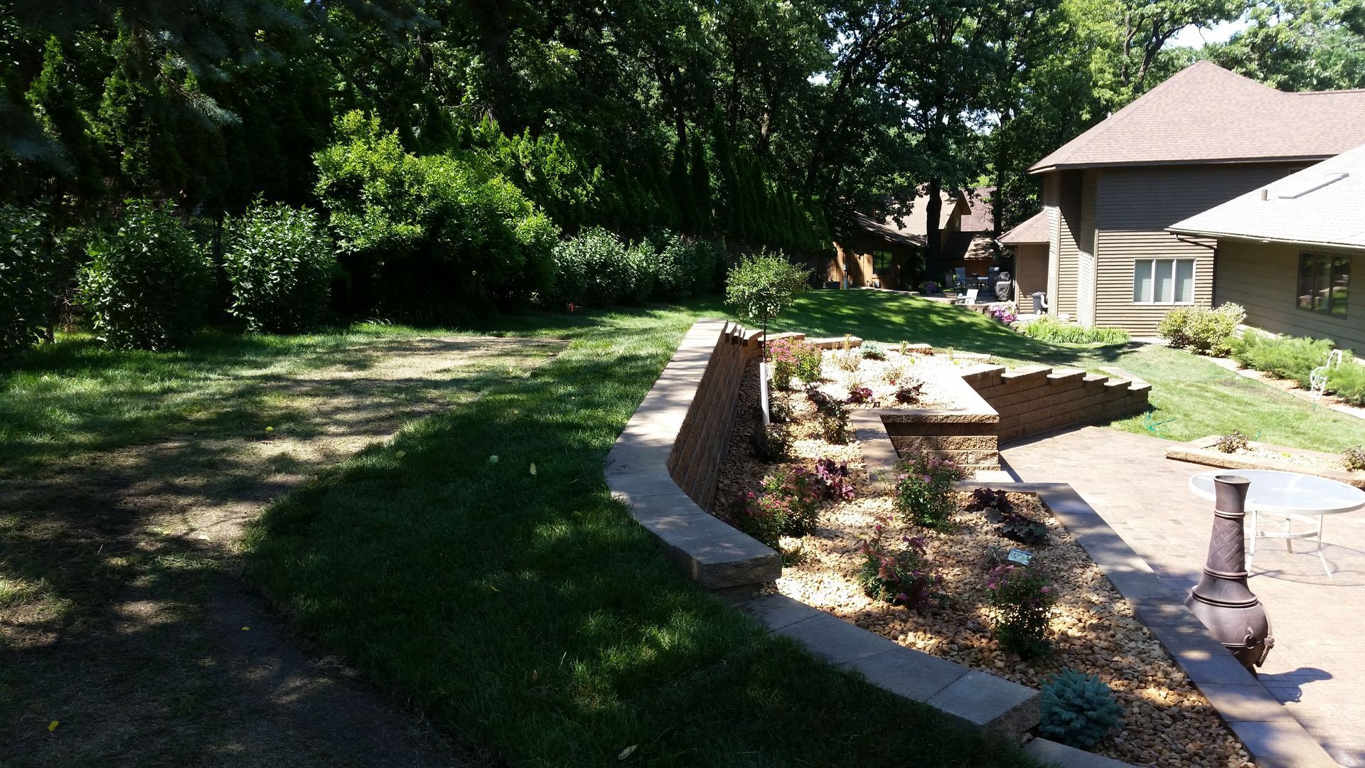 A stone retaining wall curves through a residential backyard, separating a grassy lawn from a landscaped patio area.