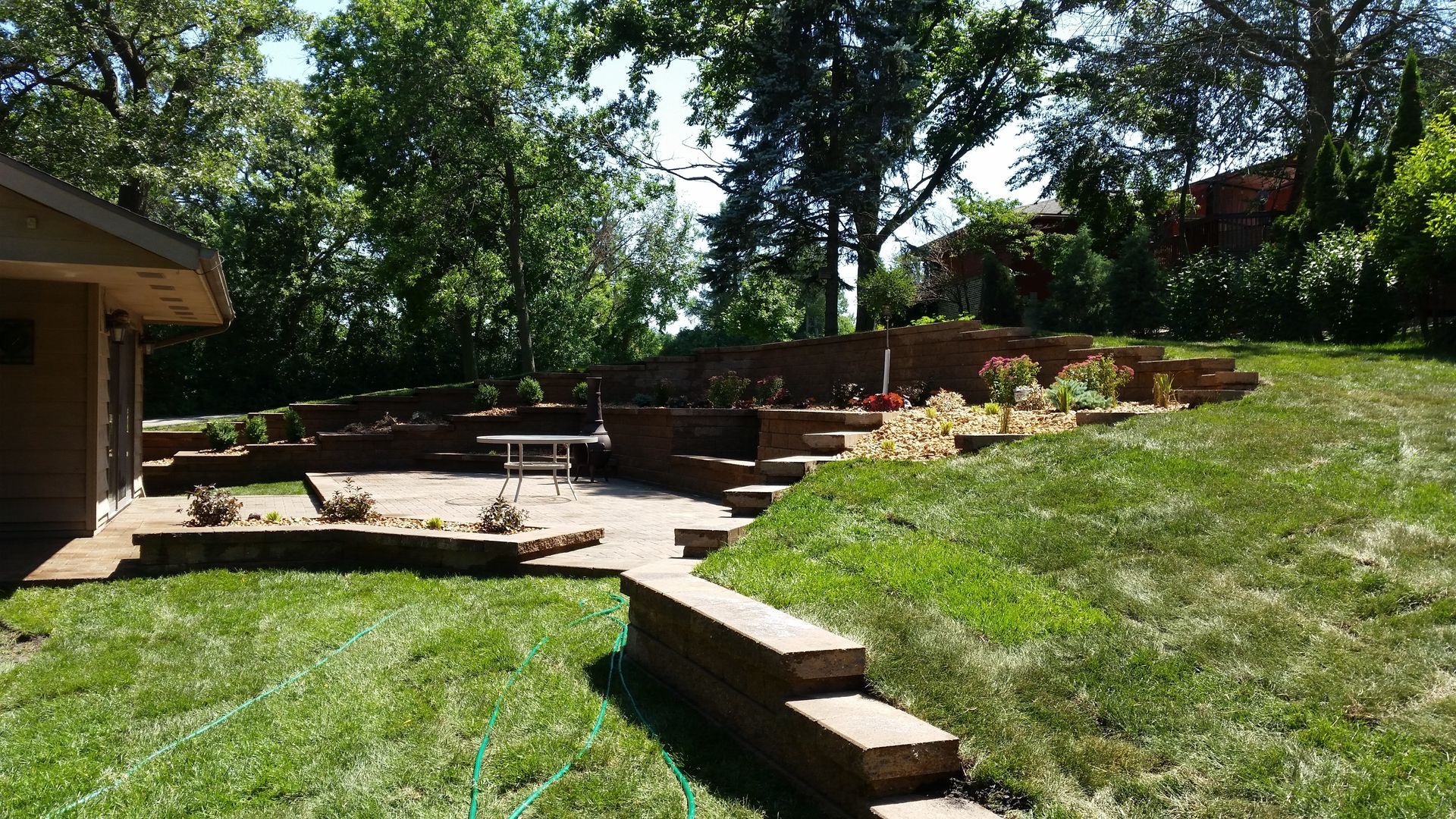 A stone patio and tiered retaining walls in a backyard, featuring a small table and landscaped beds under sunny trees.