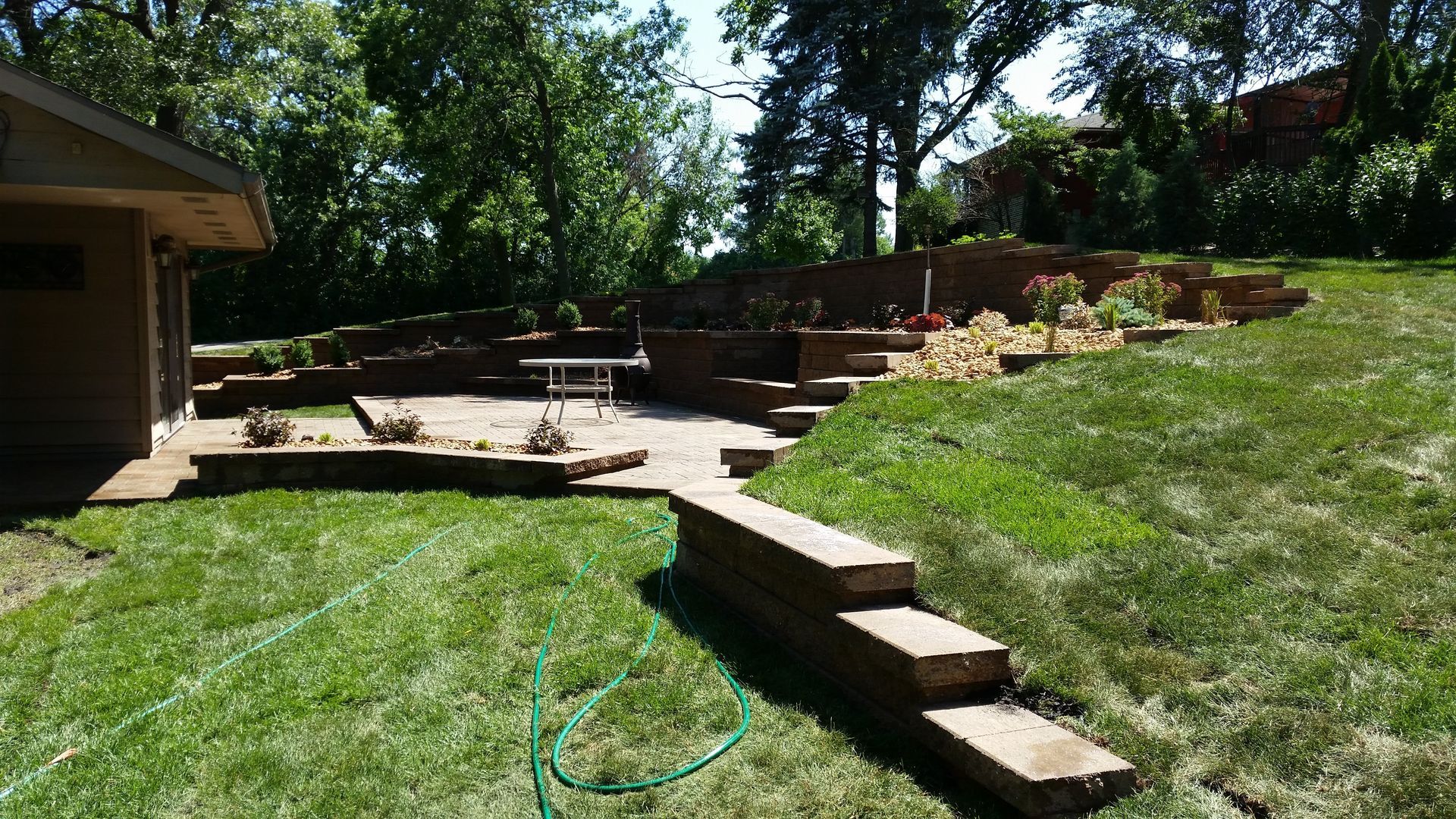 A landscaped backyard with a multi-level stone retaining wall, paved patio, and grass lawn on a sunny day.