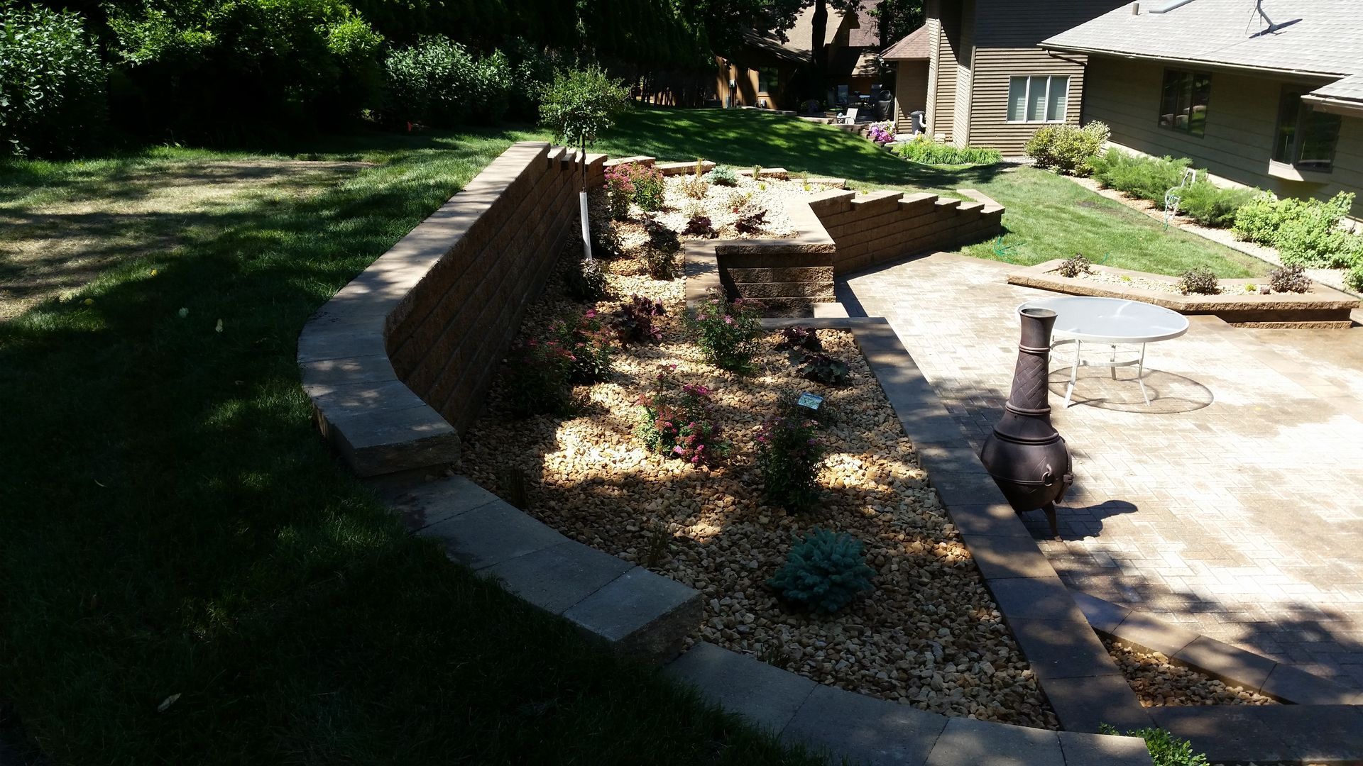 A stone retaining wall curves around a landscaped bed with gravel and shrubs next to a stone patio and a house exterior.