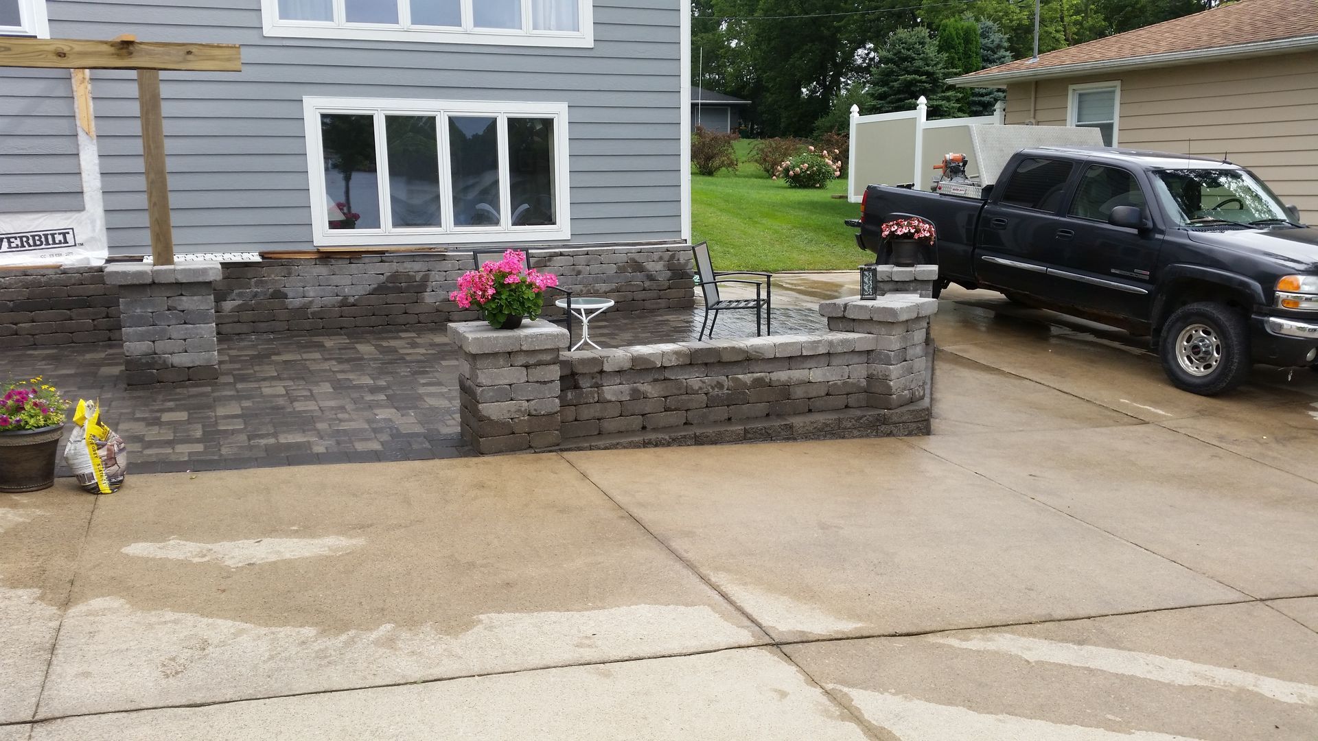 A grey house exterior with a new stone patio and low retaining wall, featuring a pickup truck parked on the driveway.