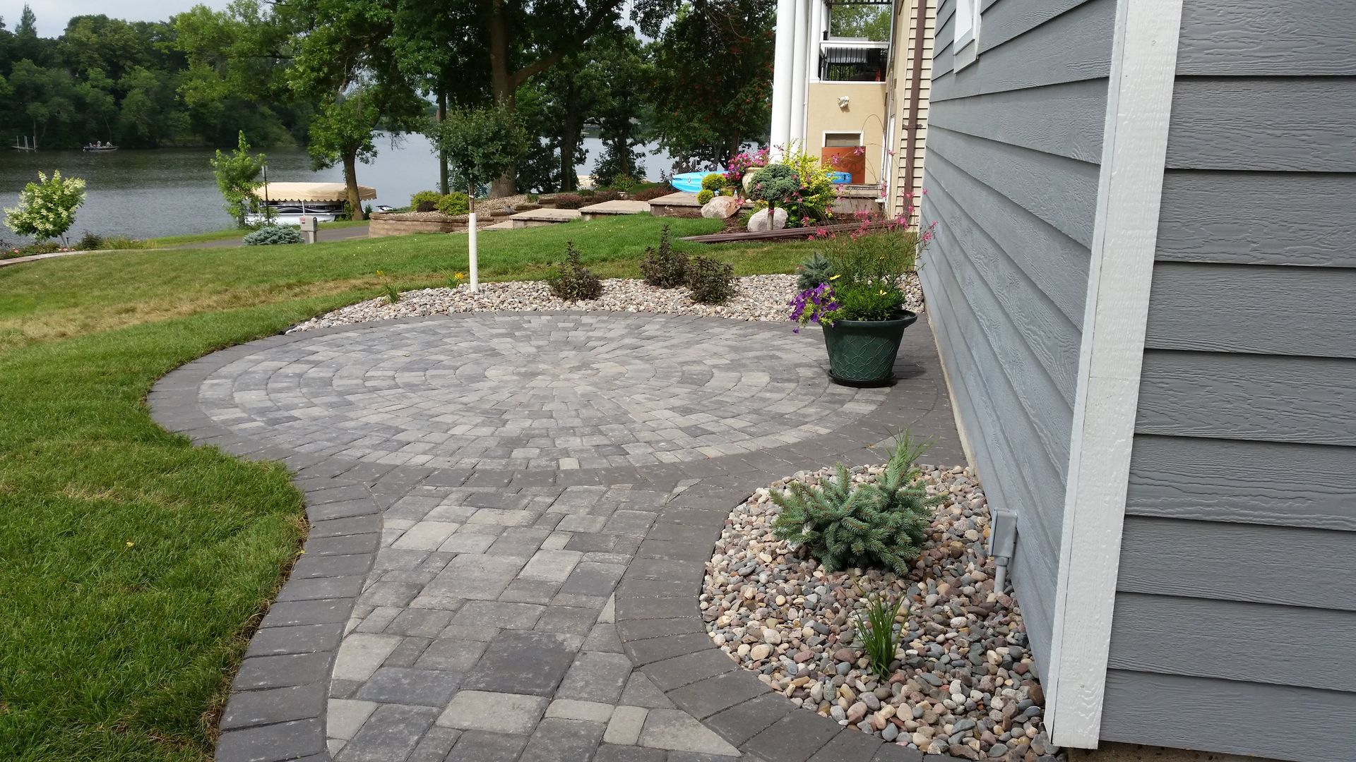 A stone patio and walkway lead to a house by a lake, surrounded by a gravel garden border and a lush green lawn.