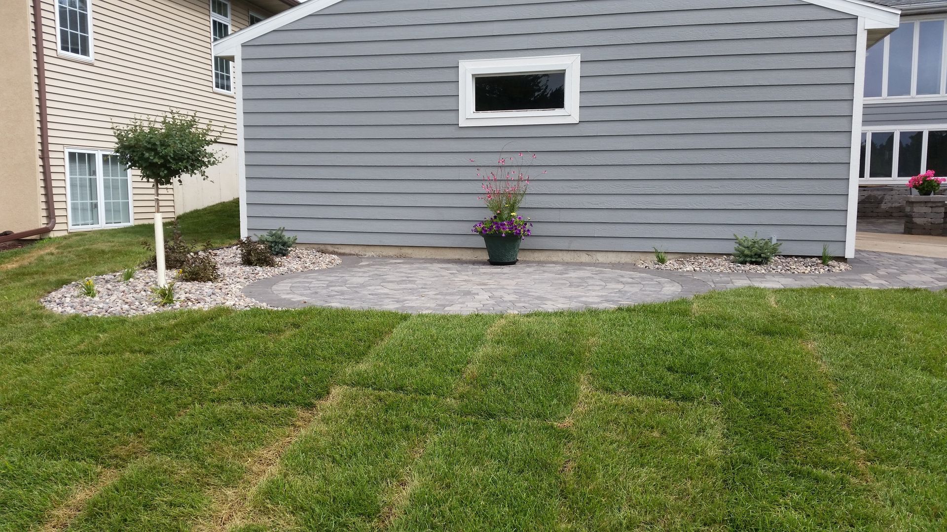 A gray-sided building with a new stone patio and landscaping, set beside a lush green lawn.