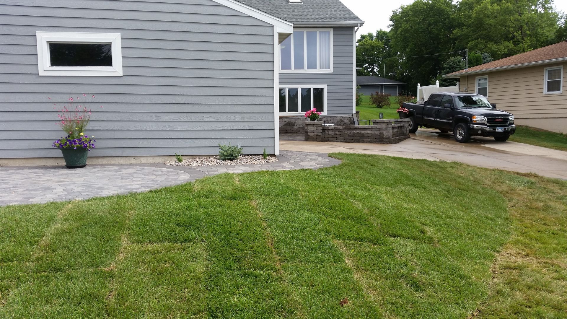 A grey-sided house with a stone patio, a small lawn in the foreground, and a black pickup truck parked in the driveway.