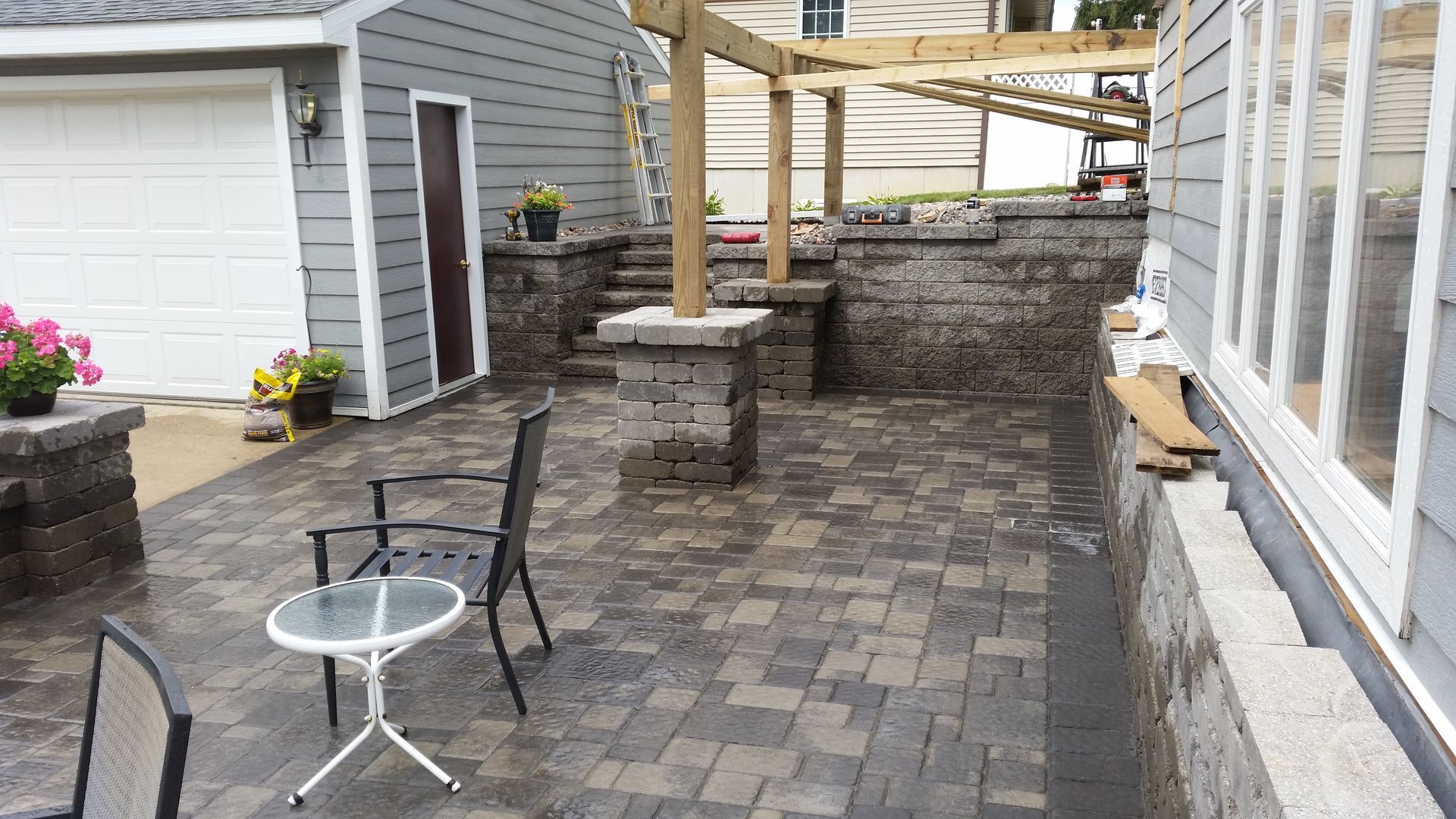 A paved outdoor patio with stone walls, a wooden pergola frame, patio chairs, and a small table next to a garage and house.
