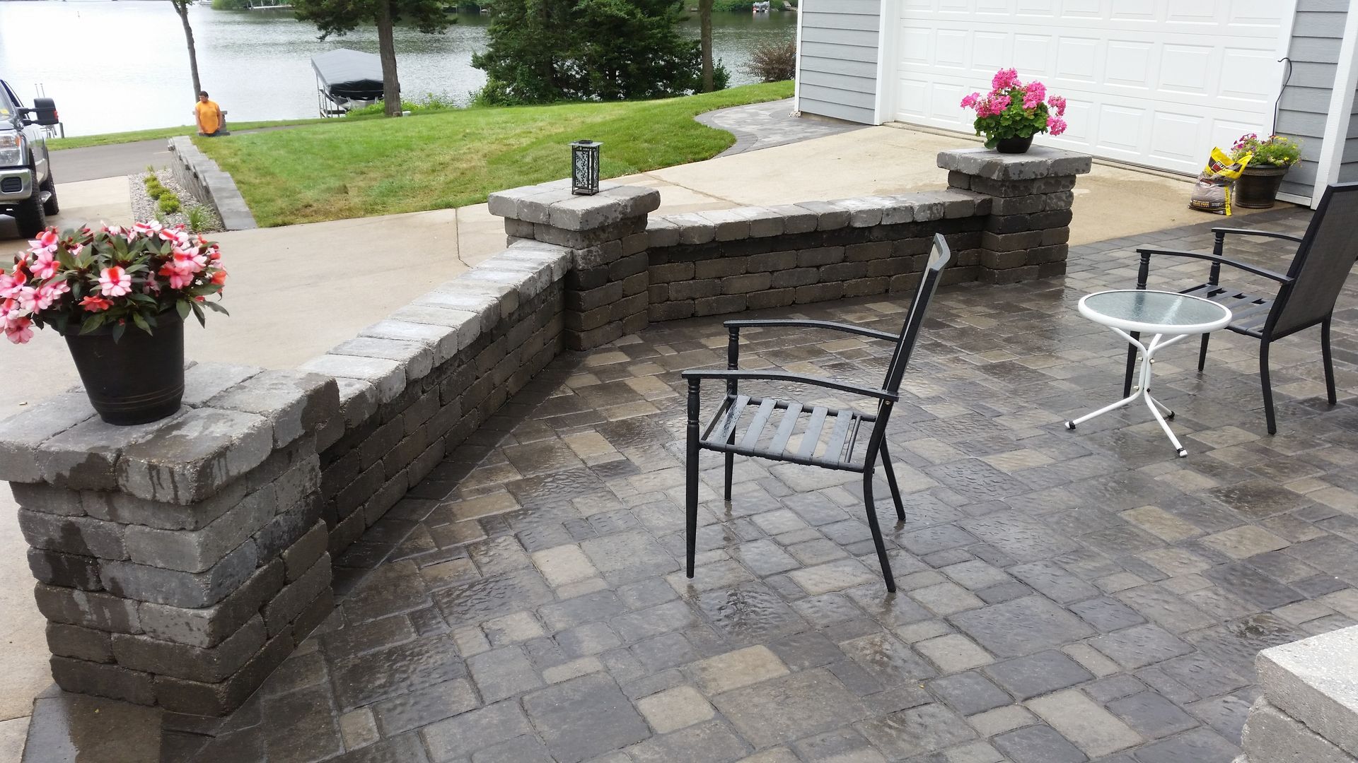 A paved patio with a curved stone retaining wall, two metal chairs, a small table, and potted pink flowers near a lake.