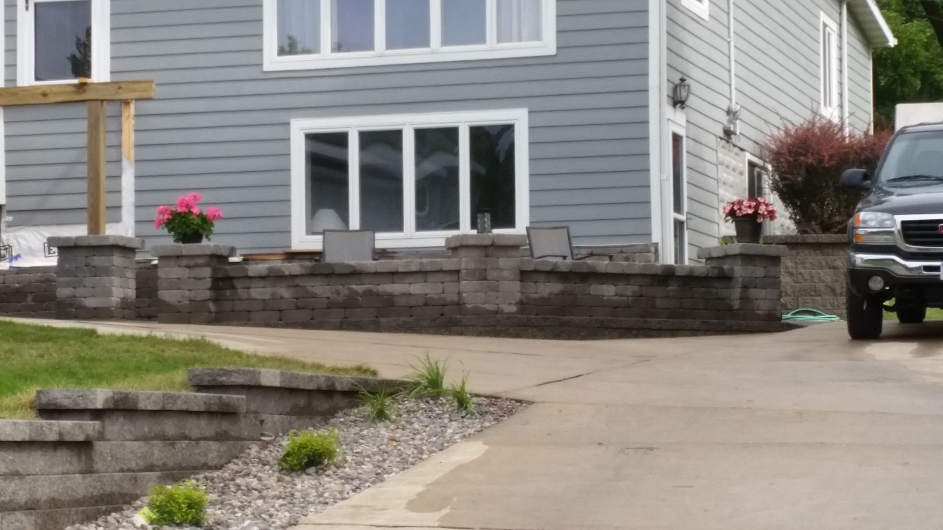 A gray house exterior with a multi-level stone retaining wall, decorative planters, and a vehicle parked on the driveway.