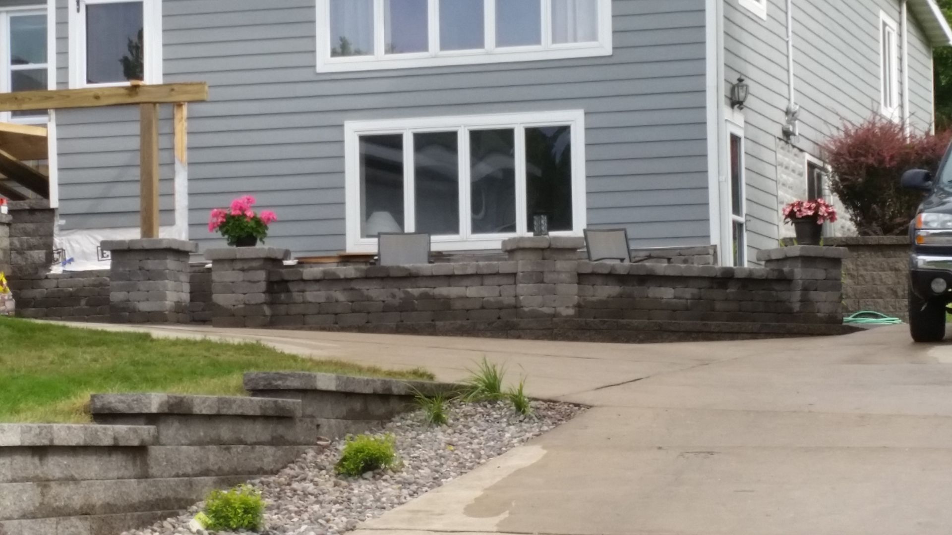 A stone retaining wall with matching pillars and a potted plant sits in front of a gray house with a concrete driveway.