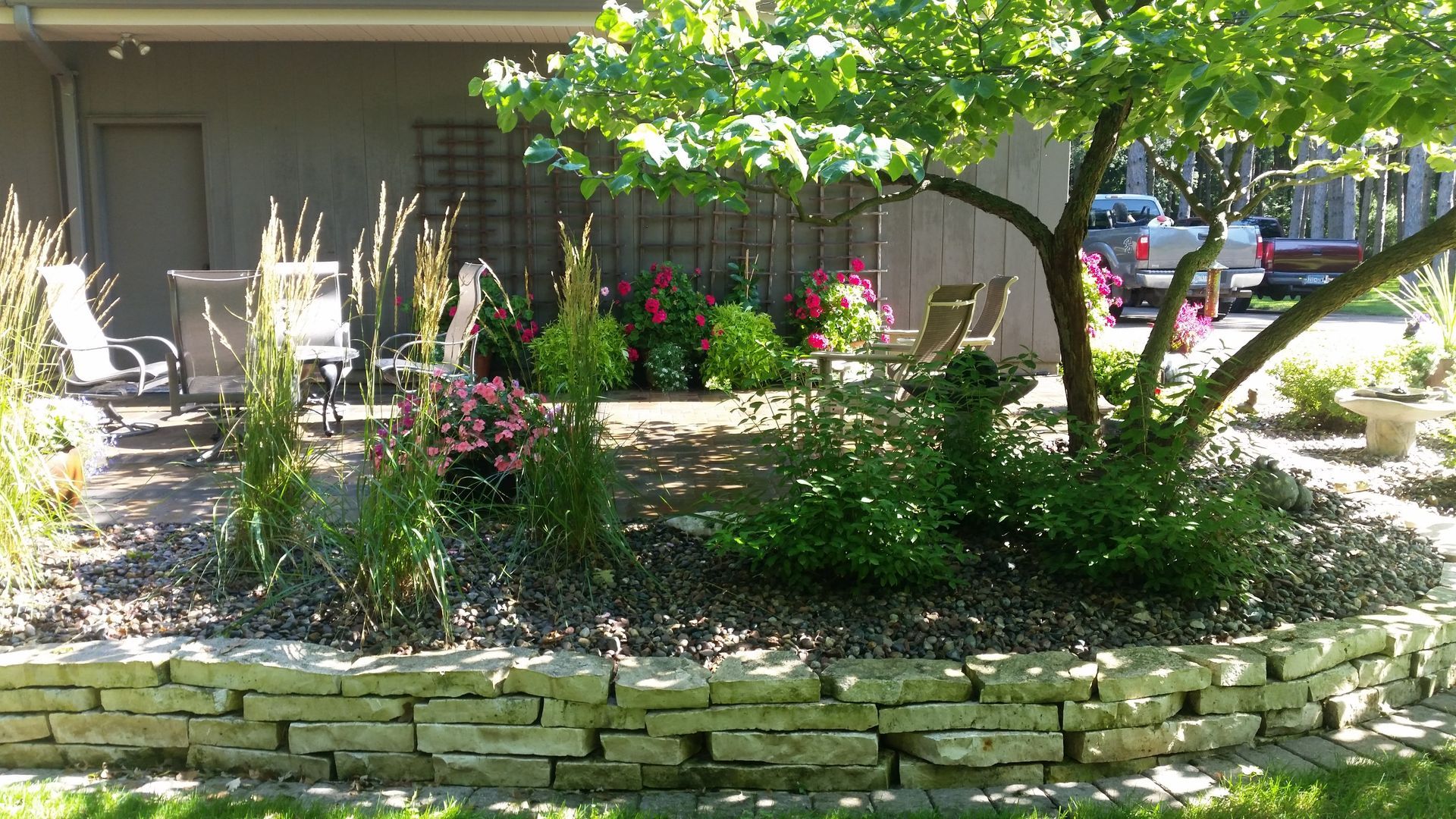 A tiered stone wall borders a garden with lush greenery, a flowering tree, and chairs set on a paved patio.
