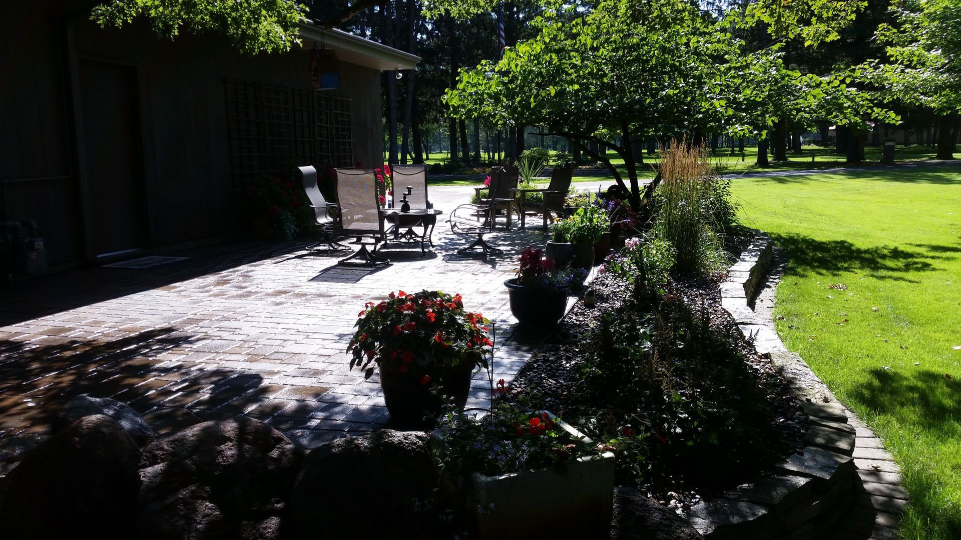 A stone patio with outdoor furniture, potted flowers, and a retaining wall, adjacent to a lush green lawn and trees.