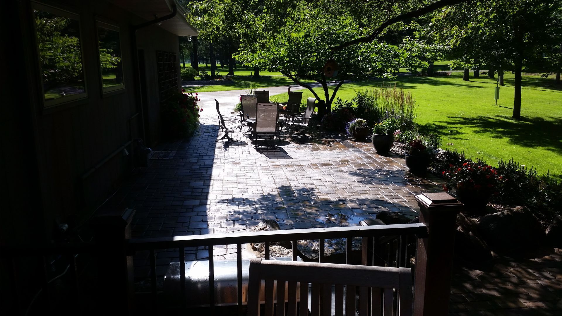 A stone patio with outdoor chairs and potted plants extends into a grassy backyard shaded by mature trees.