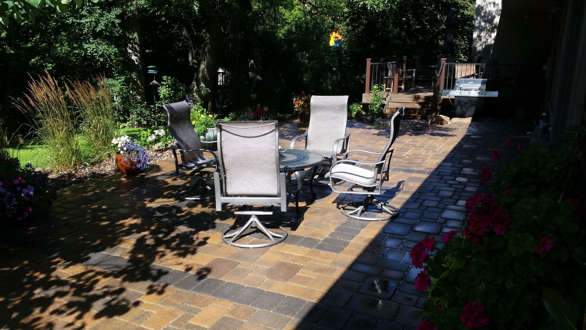 A stone patio with a table and four chairs arranged in a garden setting, illuminated by dappled sunlight.