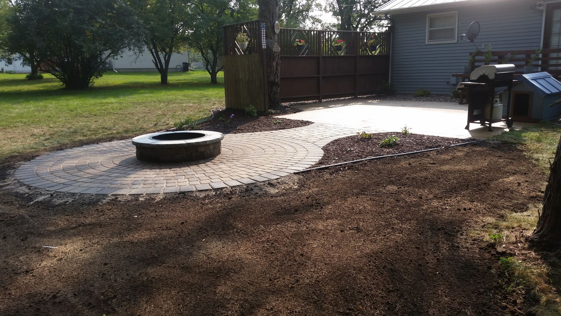 A backyard features a stone patio with a fire pit, adjacent to a wooden fence, a house wall, and a grill.