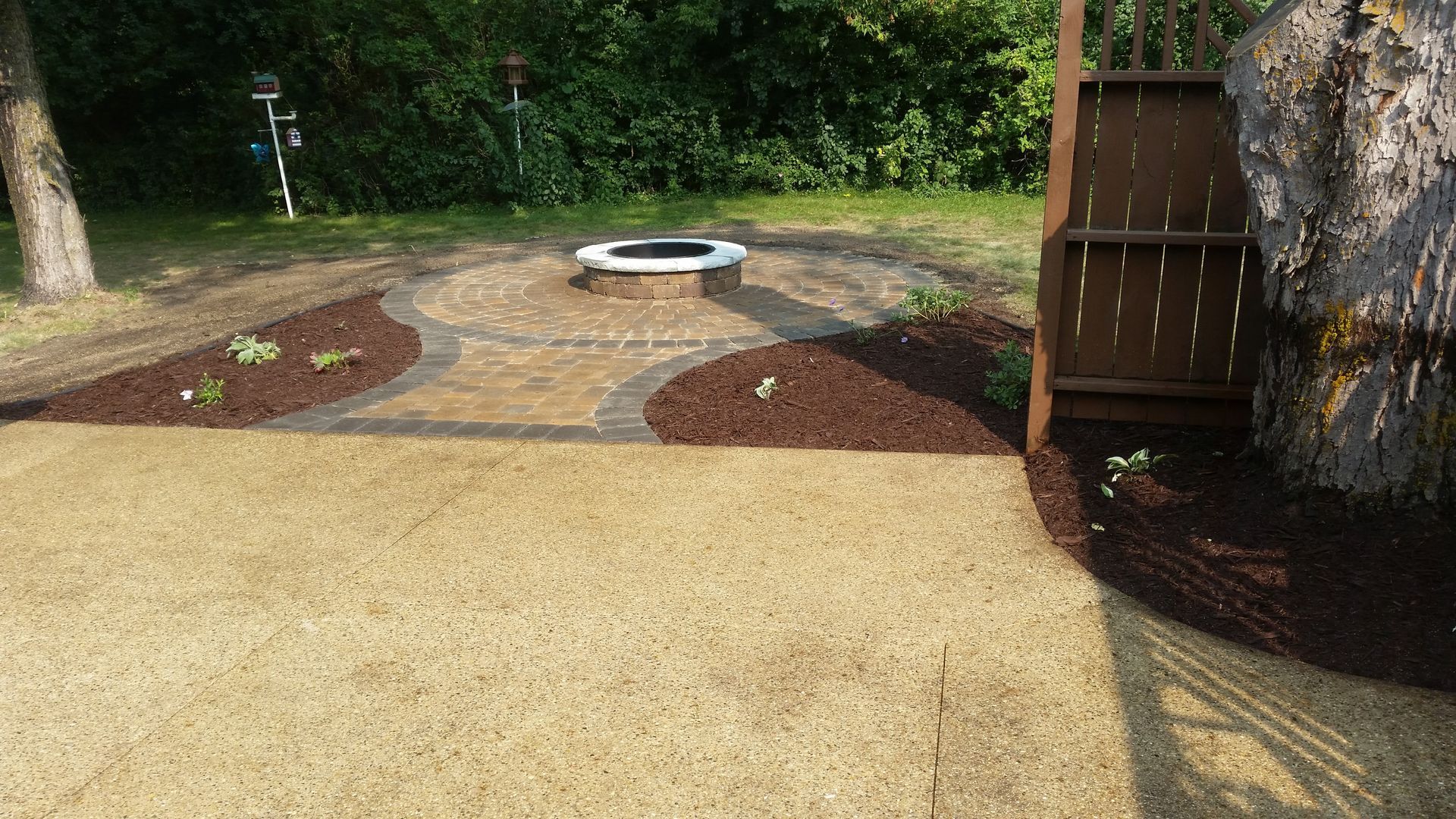 A landscaped backyard featuring a circular stone fire pit, a paved pathway, mulch garden beds, and a wooden privacy screen.