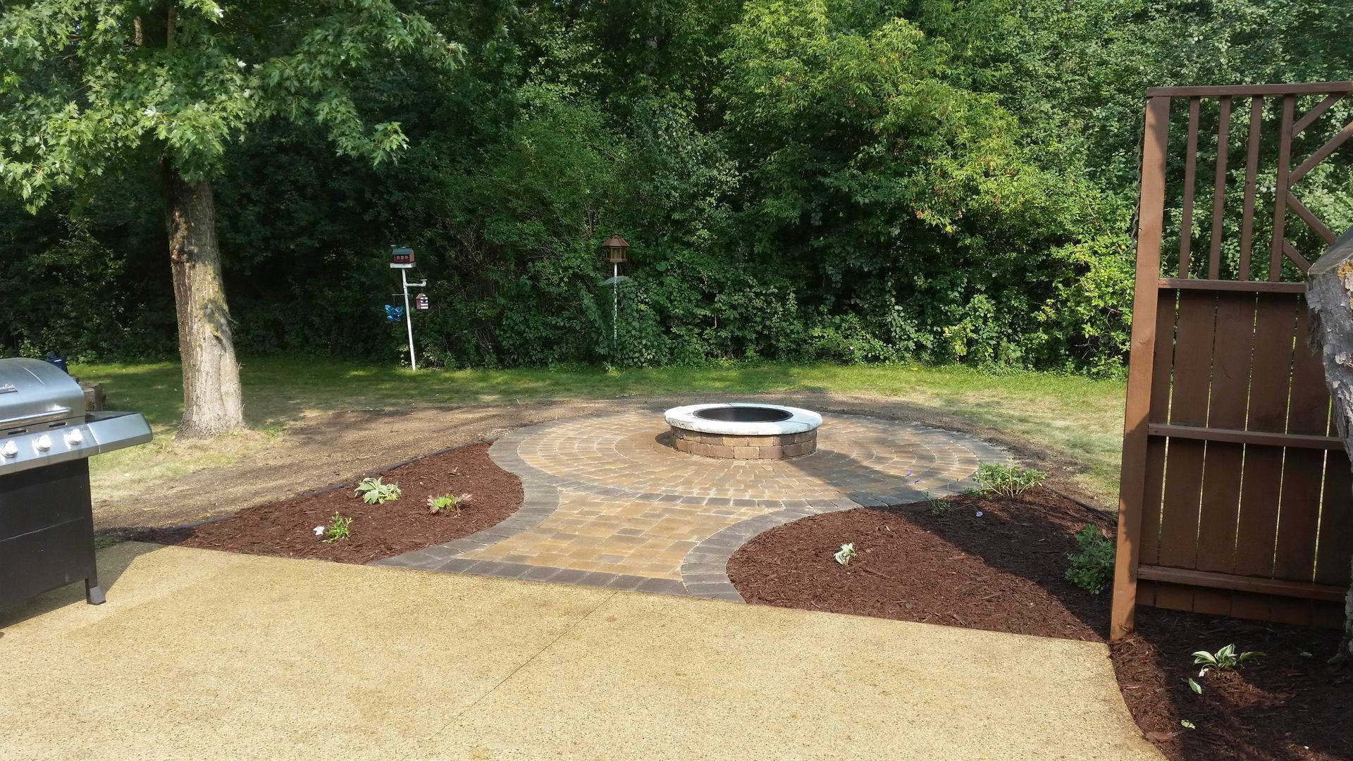 A paved patio with a circular fire pit, bordered by brown mulch beds, leading to a lawn with trees in a backyard.