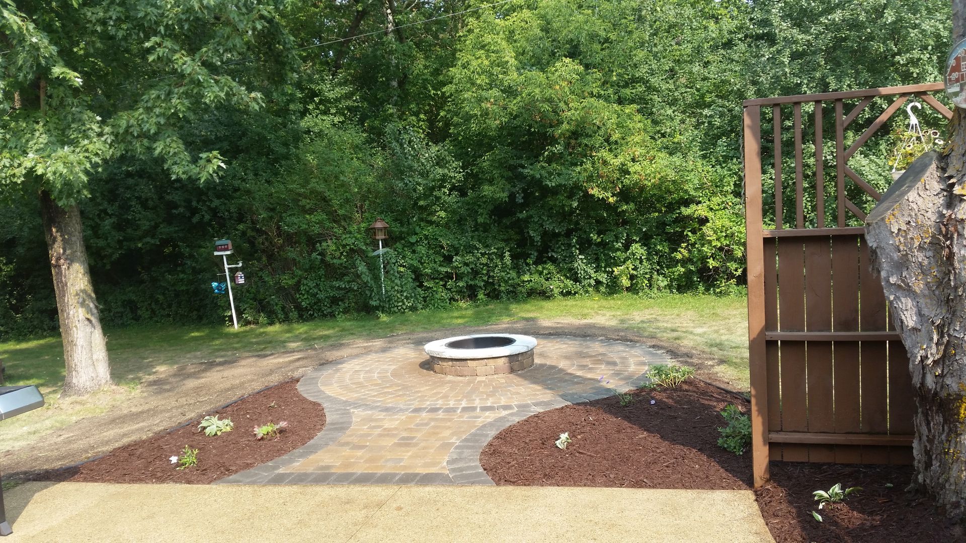 A stone fire pit centered in a circular paver patio, surrounded by dark mulch and lush green trees in a backyard setting.