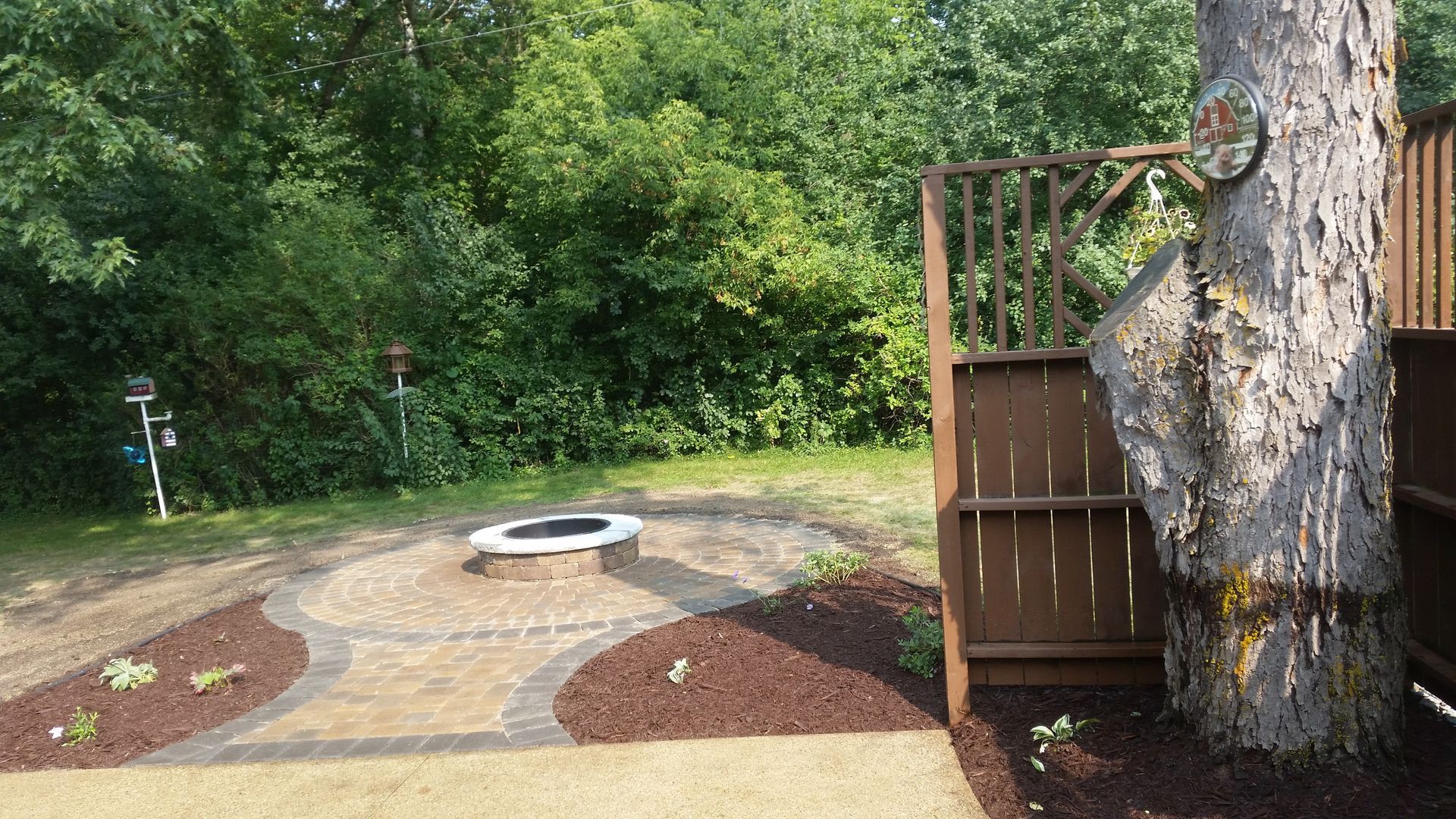 A paved circular fire pit area with stone pavers, dark mulch, and a wooden privacy screen next to a large tree.