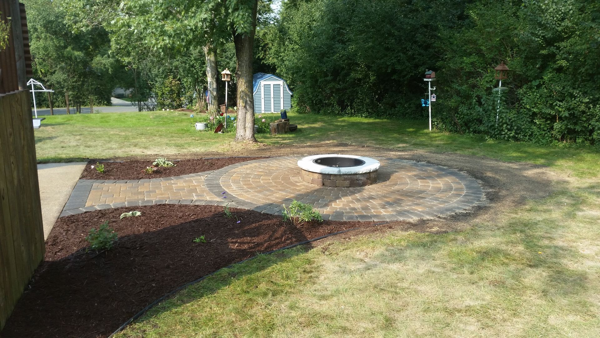 A circular brick fire pit area in a backyard, featuring a surrounding paved patio, walkway, and dark brown mulch beds.