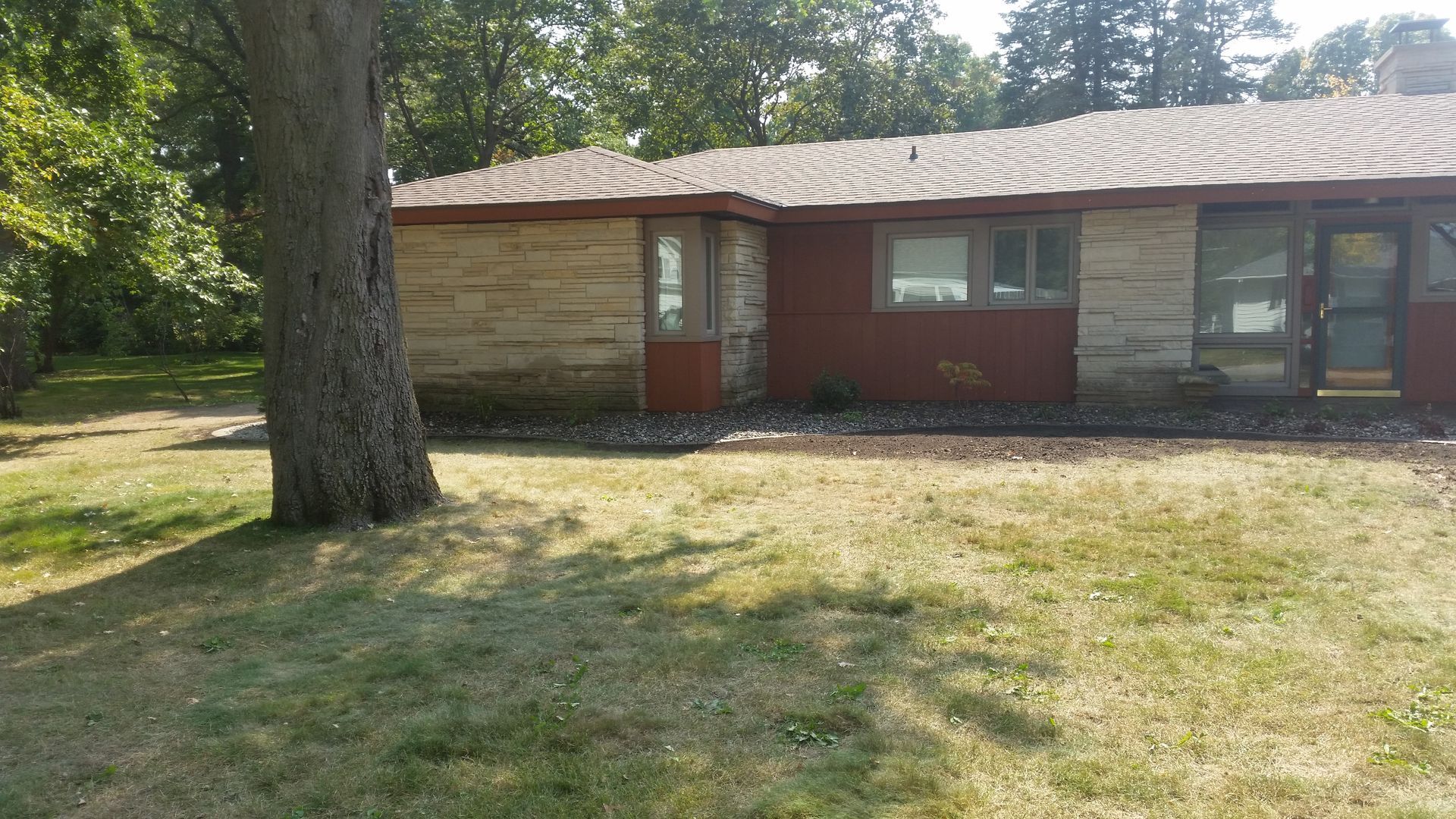 A single-story house with stone and reddish-brown siding, a brown shingled roof, and a large tree in the front yard.