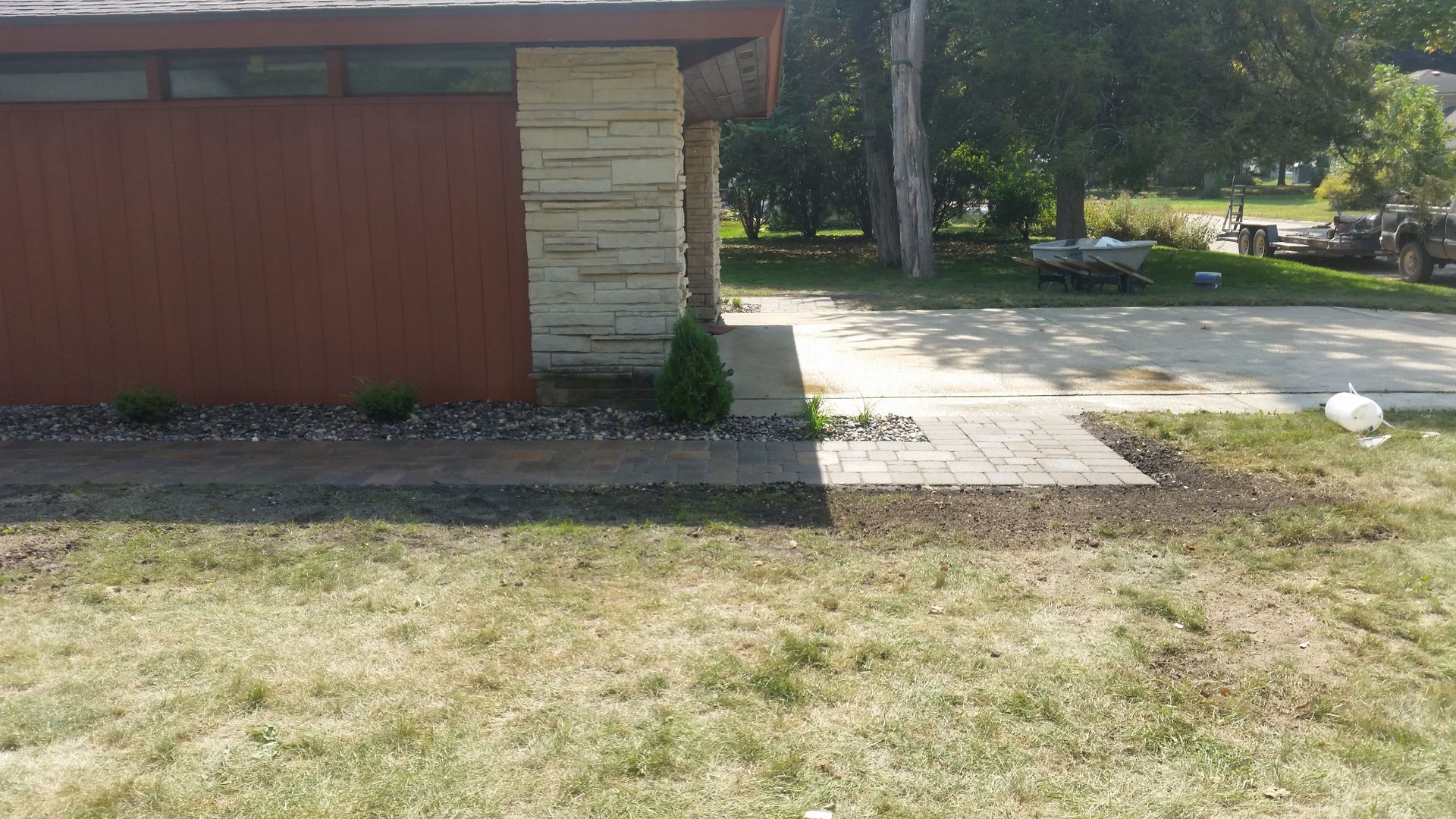 A paved walkway leads to the entrance of a house with a tan stone pillar and brown siding, next to a concrete driveway.