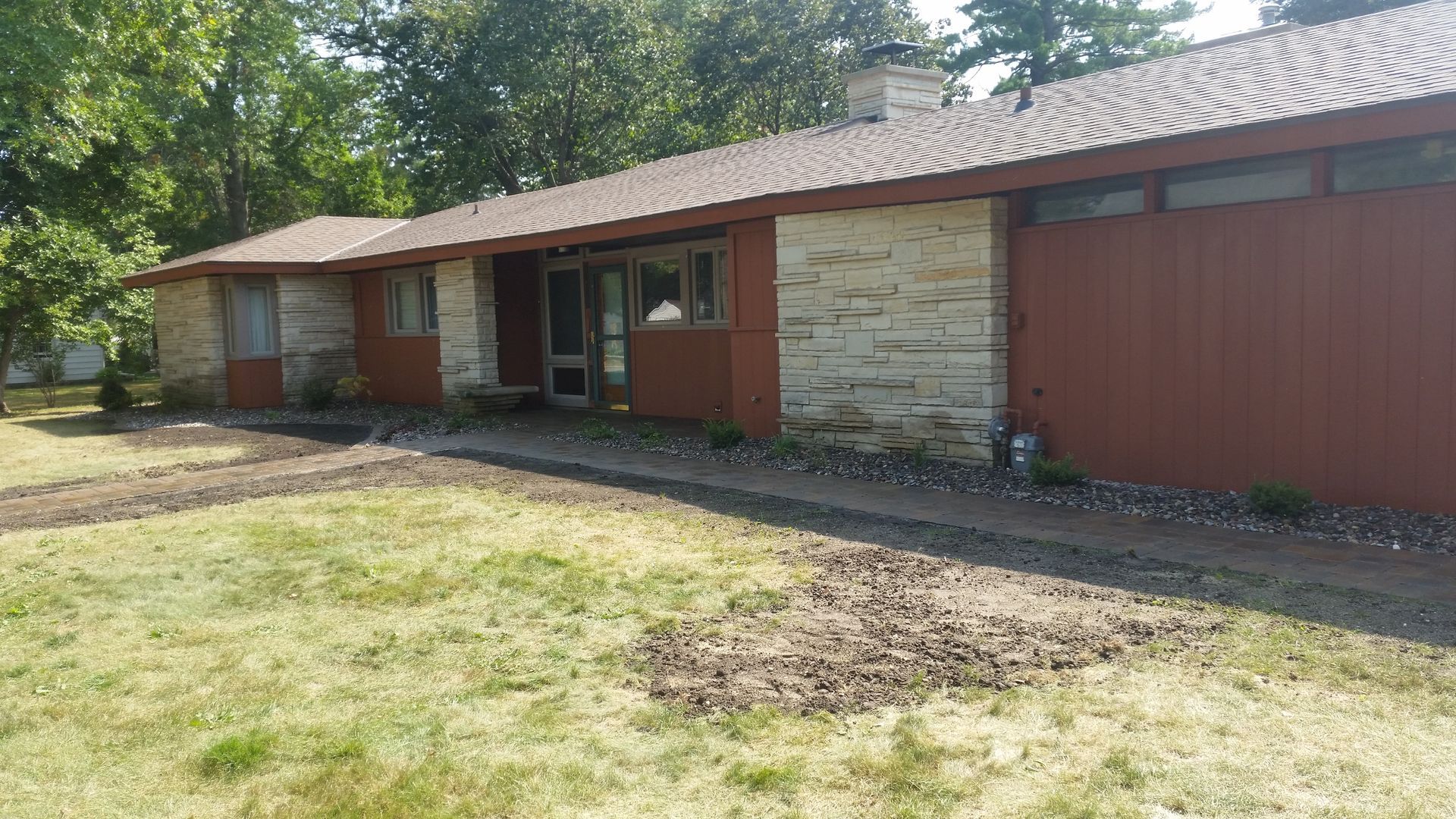 A single-story house with reddish-brown wood siding, a stone facade, and a shallow-pitched roof sits beside a grassy lawn.