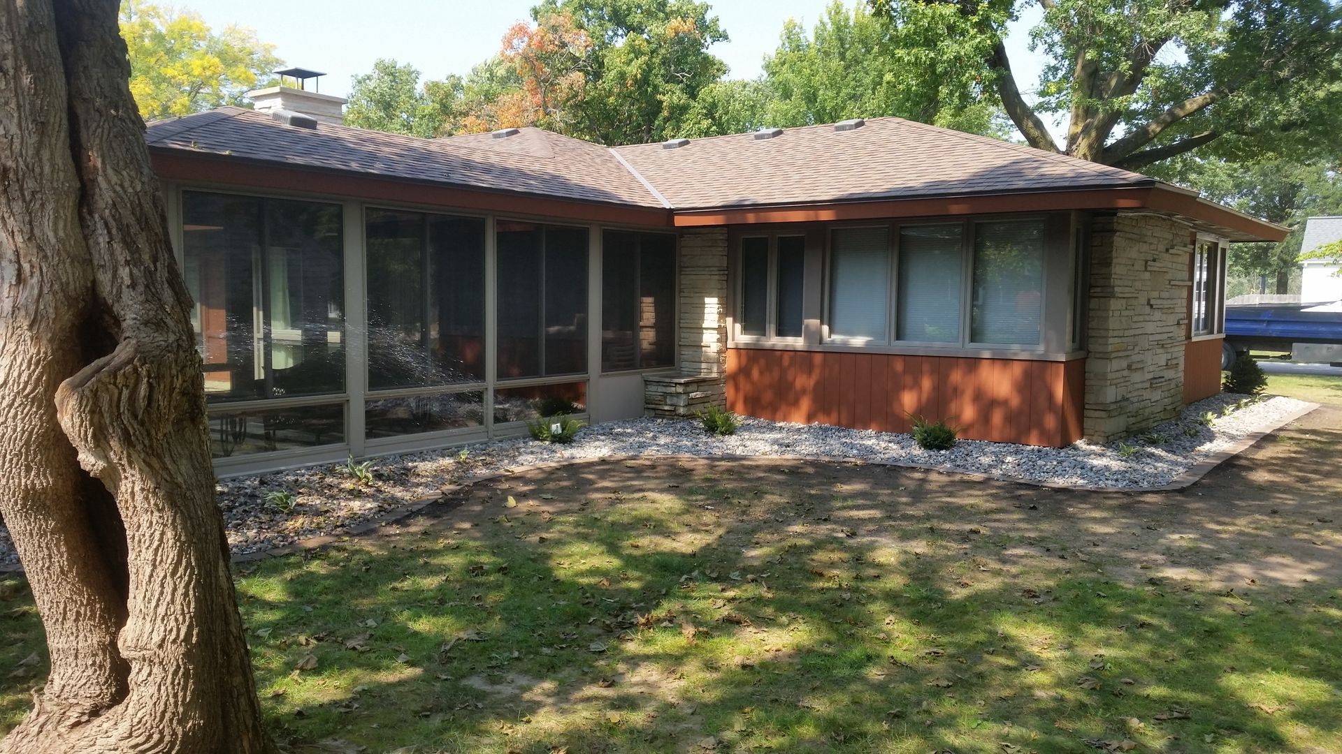 A single-story house with a screened porch and stone siding, surrounded by trees and a rocky landscape.