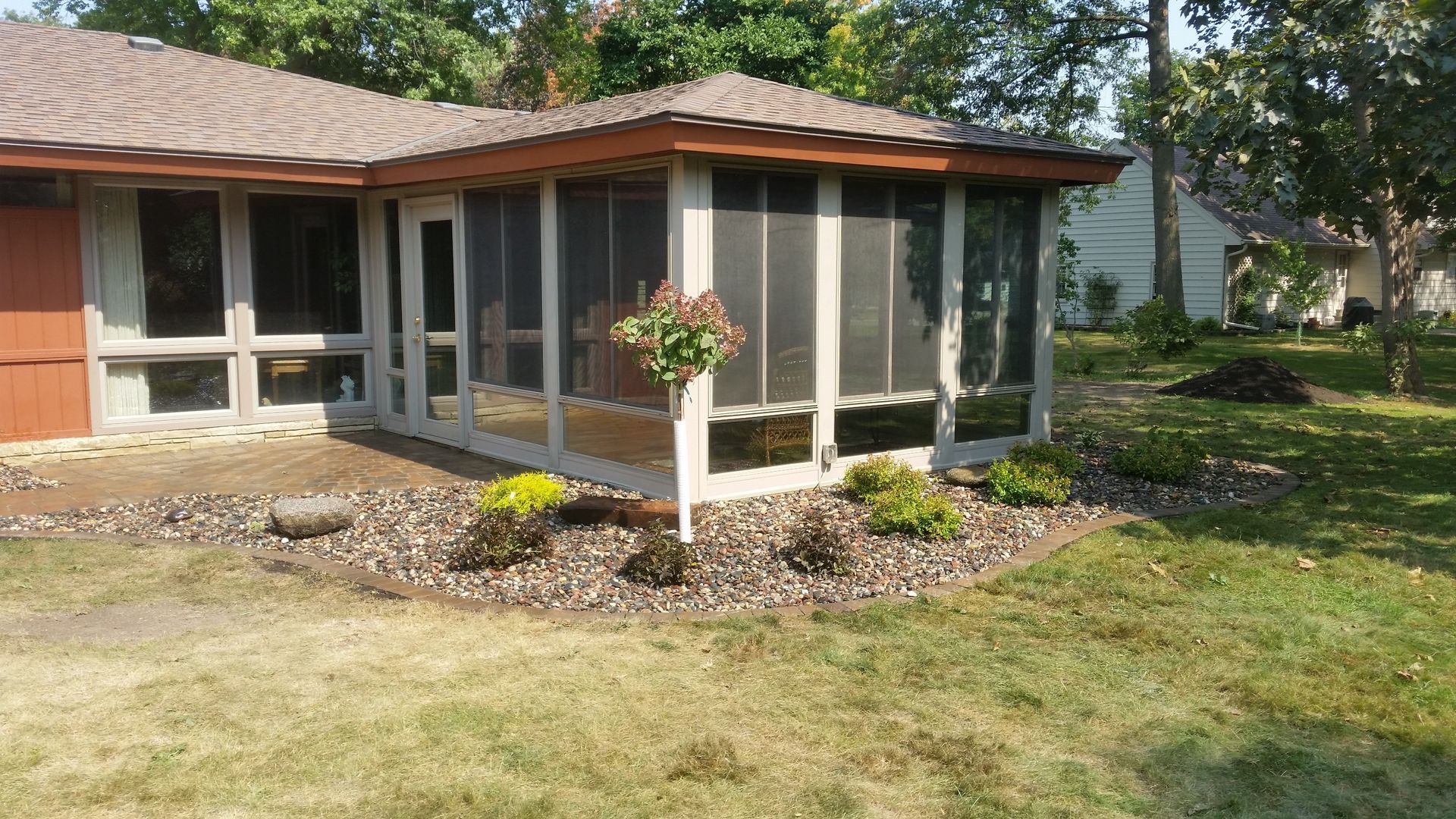 A screened-in porch addition with a brown roof stands attached to a home, surrounded by stone landscaping and lawn.