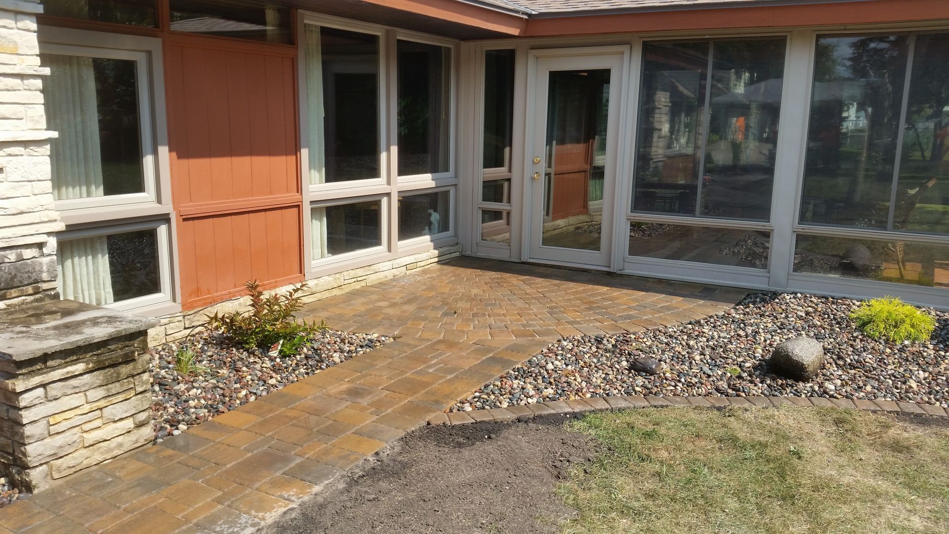 A stone walkway leads to the entrance of a house with large windows and tan siding, surrounded by a gravel garden bed.