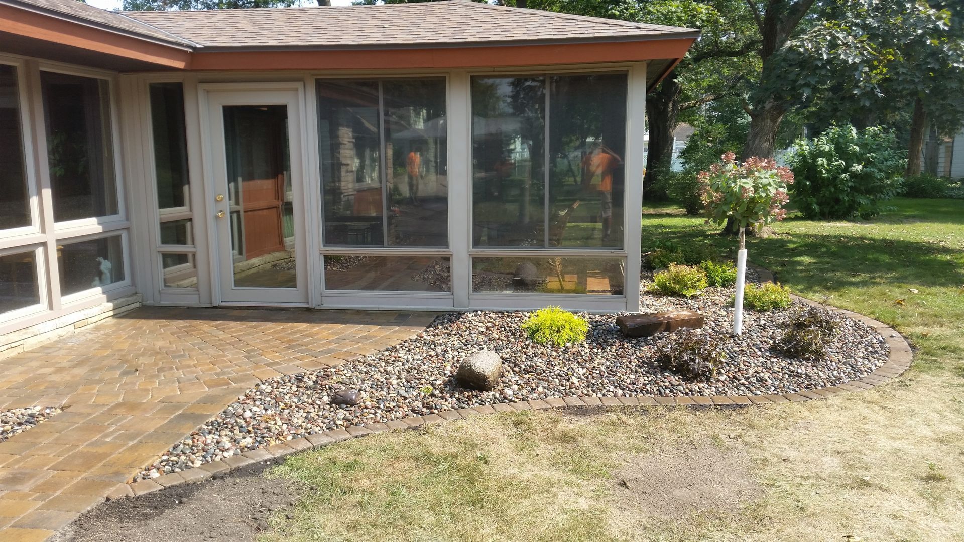 A sunroom with a white door next to a brown patio and a landscaped rock garden with a small tree on a grassy lawn.