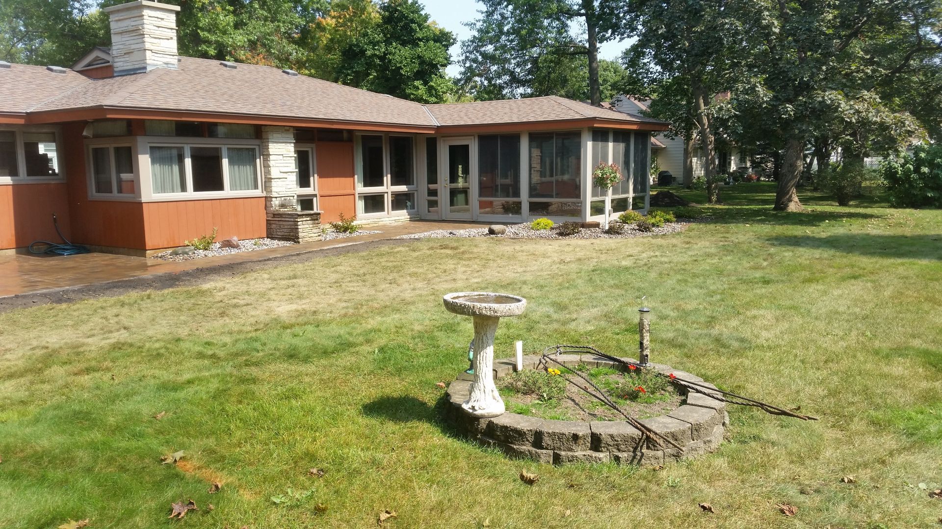 A tan house with a stone chimney and a screened porch sits behind a circular stone garden bed with a birdbath.