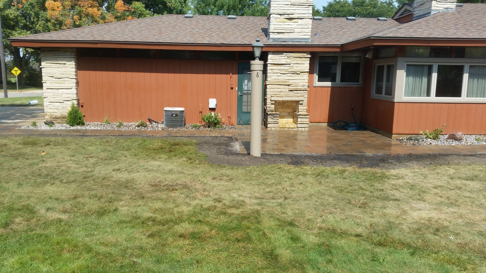A ranch-style house with brown siding, stone accents, a stone chimney, and a paved patio area under a sunny sky.