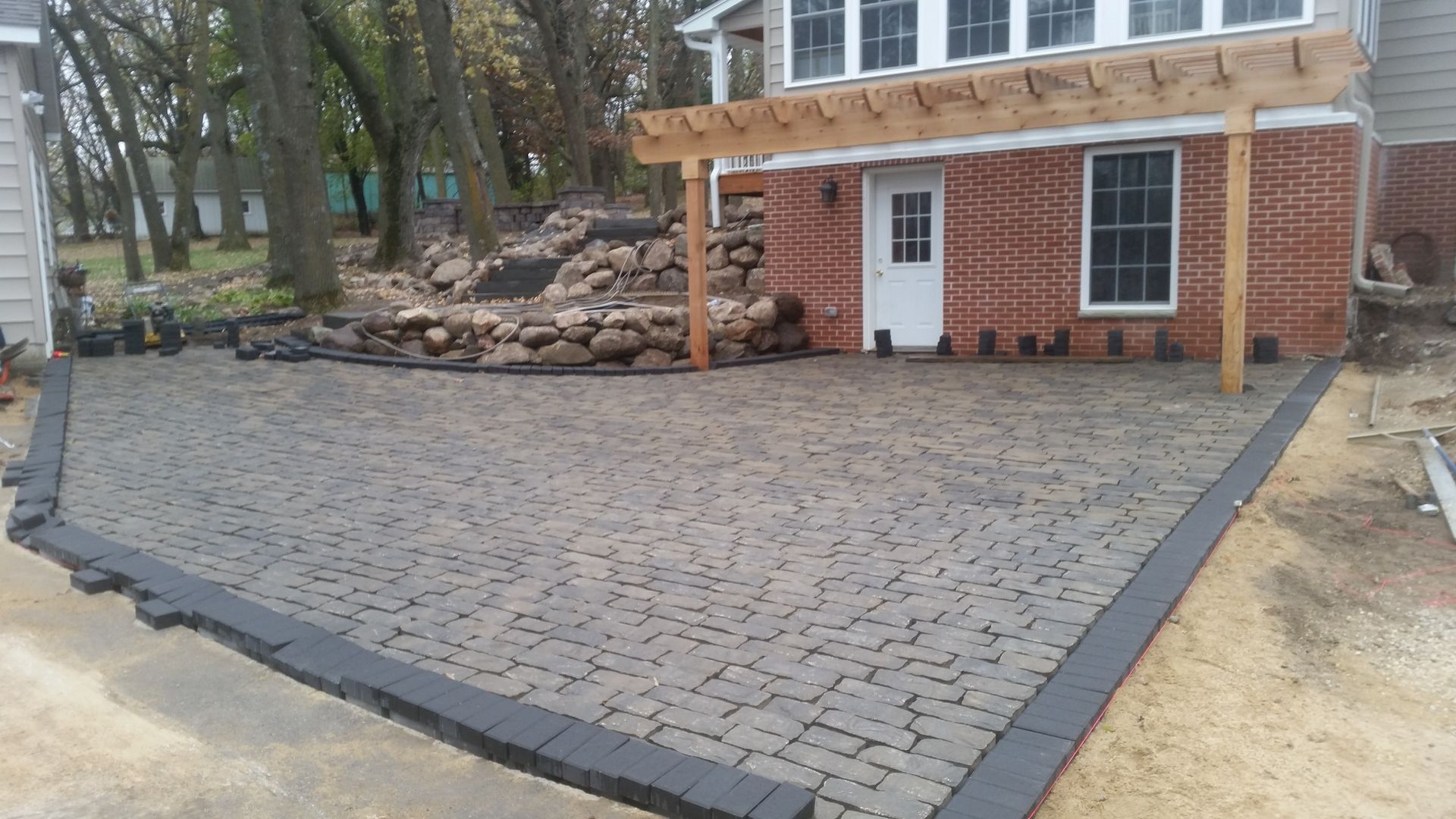 A newly installed gray paver patio with a dark border sits in front of a brick house with a wooden pergola.