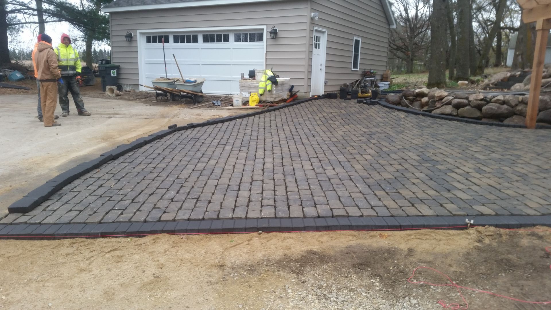Two construction workers stand near a newly laid gray paver stone patio in front of a tan garage.