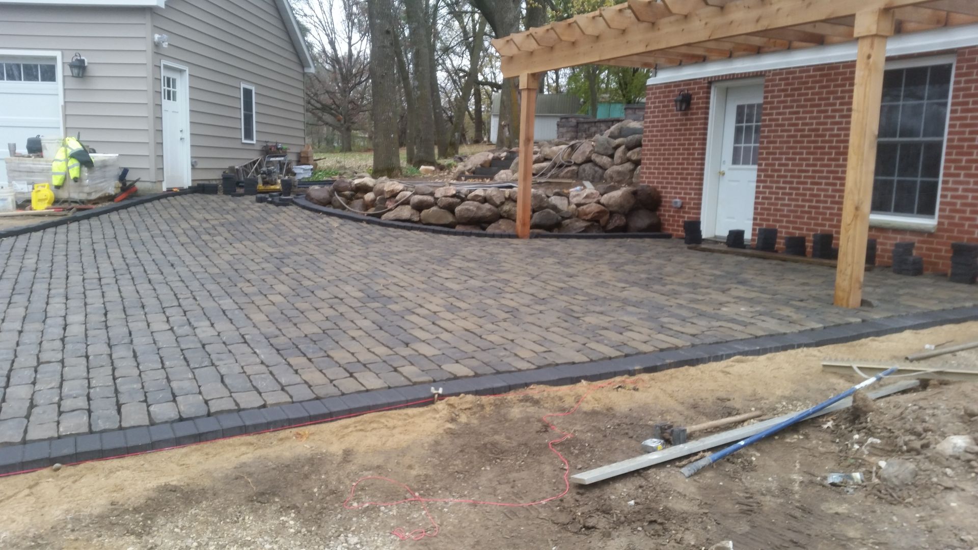 A newly installed gray paver patio with a dark border and a wooden pergola attached to a brick house.