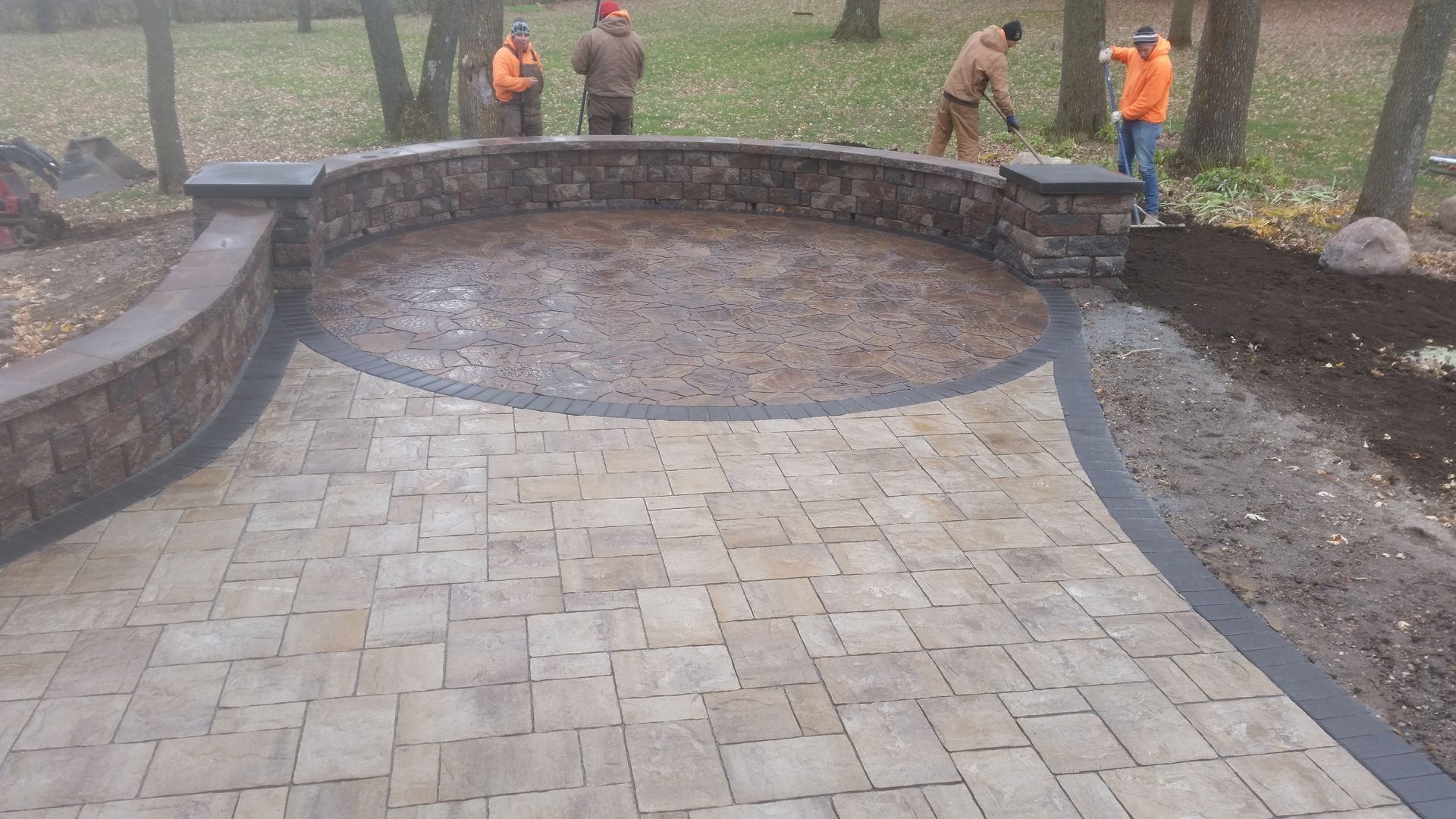 Construction workers building a curved stone wall and paver patio in a wooded yard.
