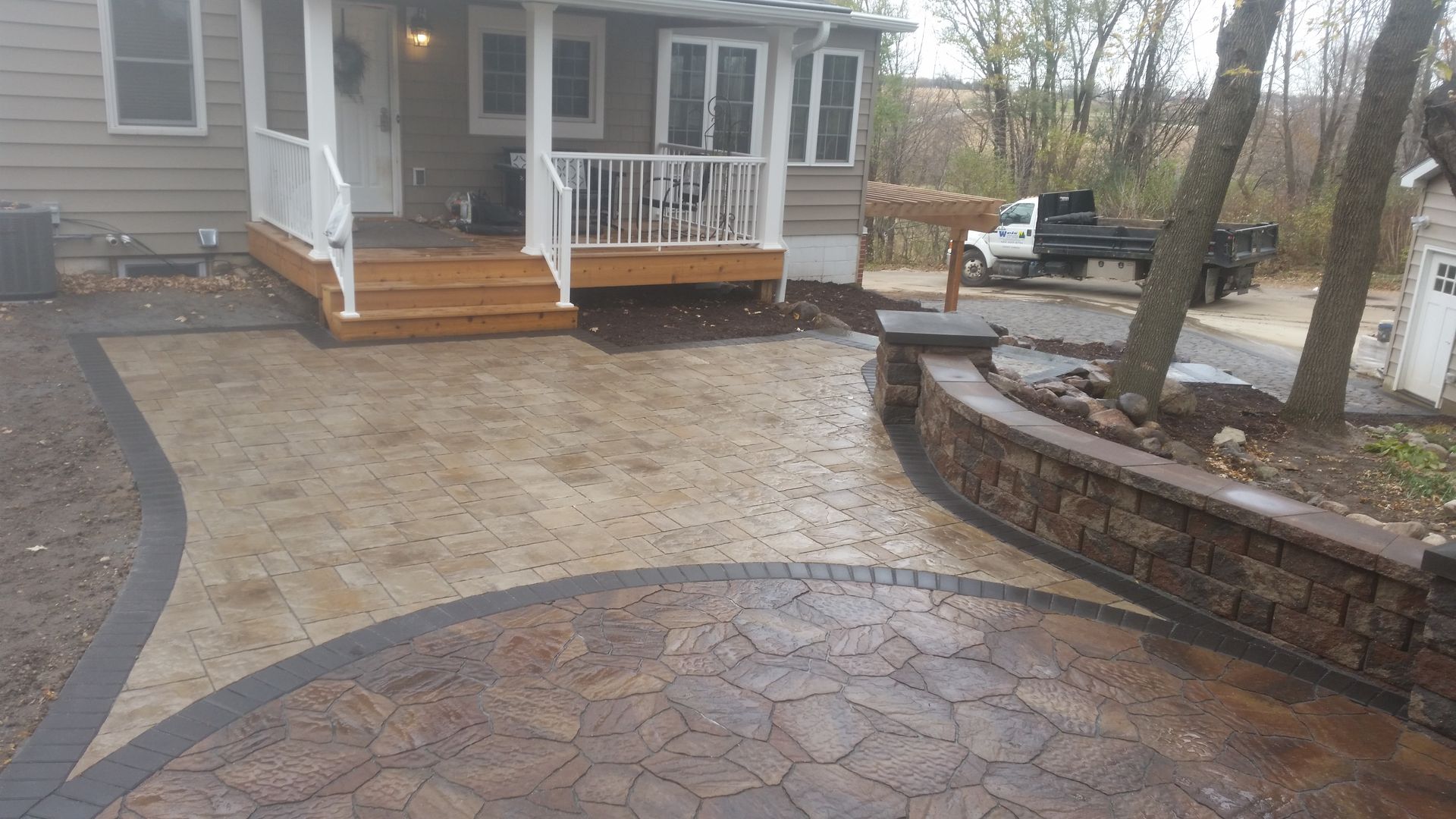 A tan and brown paver patio with a curved stone retaining wall leads to wooden steps and a white porch on a house.