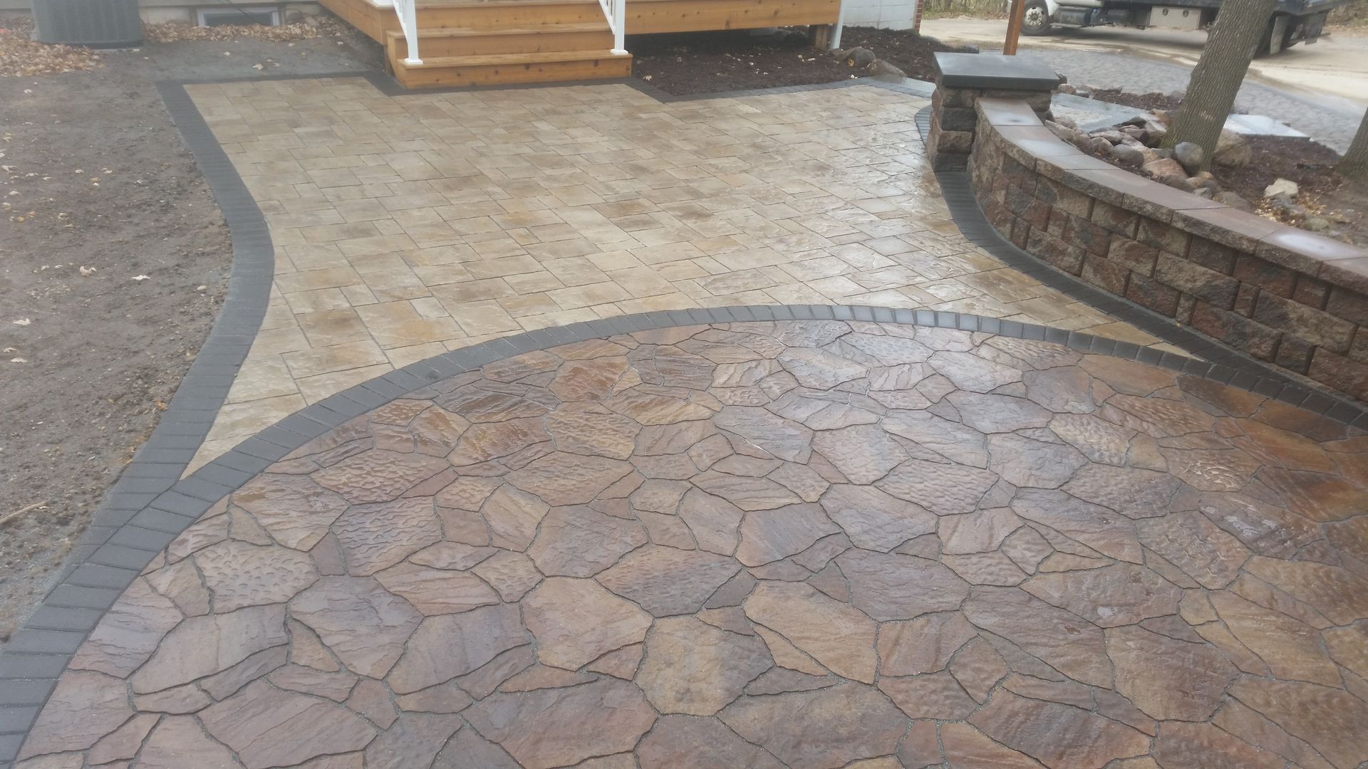 A stone patio featuring two contrasting patterns, with a curved retaining wall and wooden steps in the background.