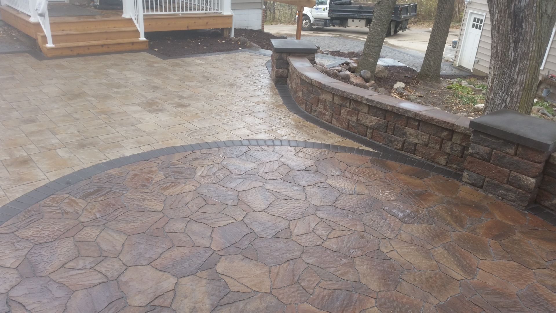 A patio with irregular brown stone pavers, a curved stone retaining wall, and a staircase leading to a white deck.